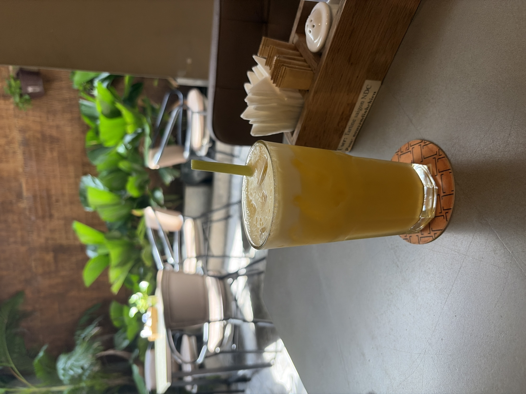 The image depicts a static, eye-level view of a light-colored table in an outdoor or semi-outdoor cafe in Thủ Đức, Vietnam, specifically an establishment identified as "Tartine Saigon NDC" by text on a wooden caddy. In the foreground, a tall, clear glass contains a vibrant yellow beverage topped with a frothy white layer, and a bright green straw is inserted. The glass sits on a round, orange-patterned coaster. To the right, a rectangular wooden caddy holds a fan of white folded paper napkins, multiple individual brown sweetener packets arranged vertically, and a small white ceramic salt shaker. A white label on the caddy displays "User: Tartine saigon NDC" and "Pass: nguyendinhhieuvn". The midground and background are soft-focused, revealing a lush green garden setting with various large-leafed plants. A wooden plank wall serves as a backdrop, with a small potted plant hanging on it in the upper right. Blurry outlines of light-colored tables and dark-framed chairs are visible among the foliage, indicating additional seating areas. No individuals are present.