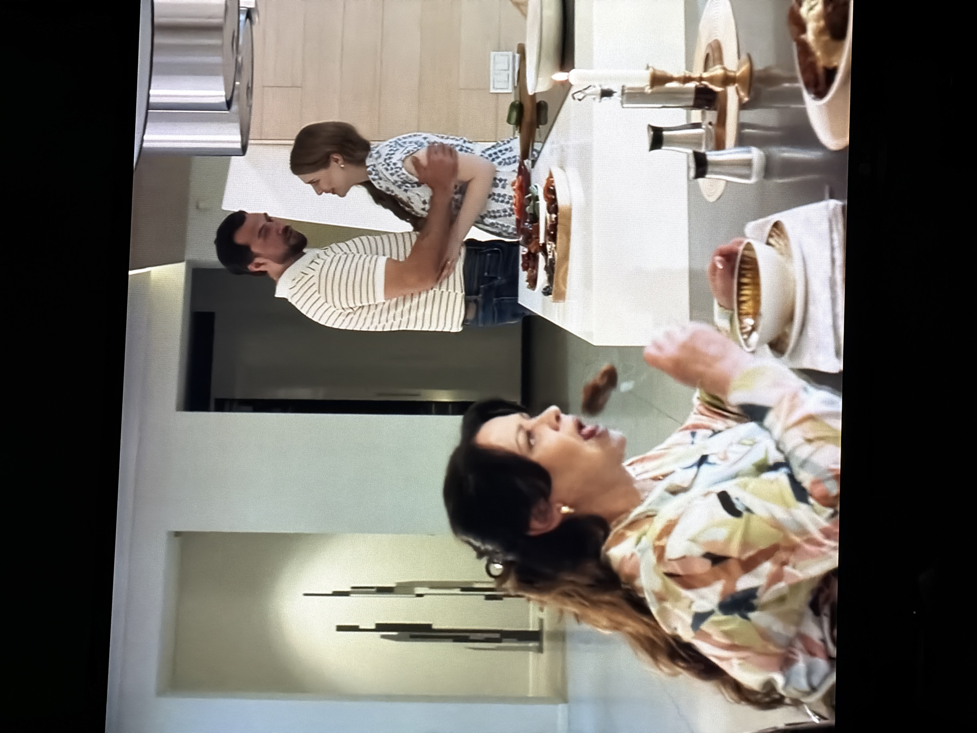 The image shows an indoor scene, likely a modern kitchen and living area. In the foreground, a woman with dark hair pulled back in a ponytail is seated at a table, holding a fork with food towards her mouth. She is wearing a colorful, patterned shirt. In the background, a man and a woman are embracing. The man is wearing a striped shirt and jeans, and the woman is wearing a patterned dress. The environment appears to be a well-lit, contemporary home with light-colored walls, modern fixtures, and decorative elements. There are serving dishes with food on the counter. The location is not in the city of Semiluki, Russia; it is an indoor residential setting.