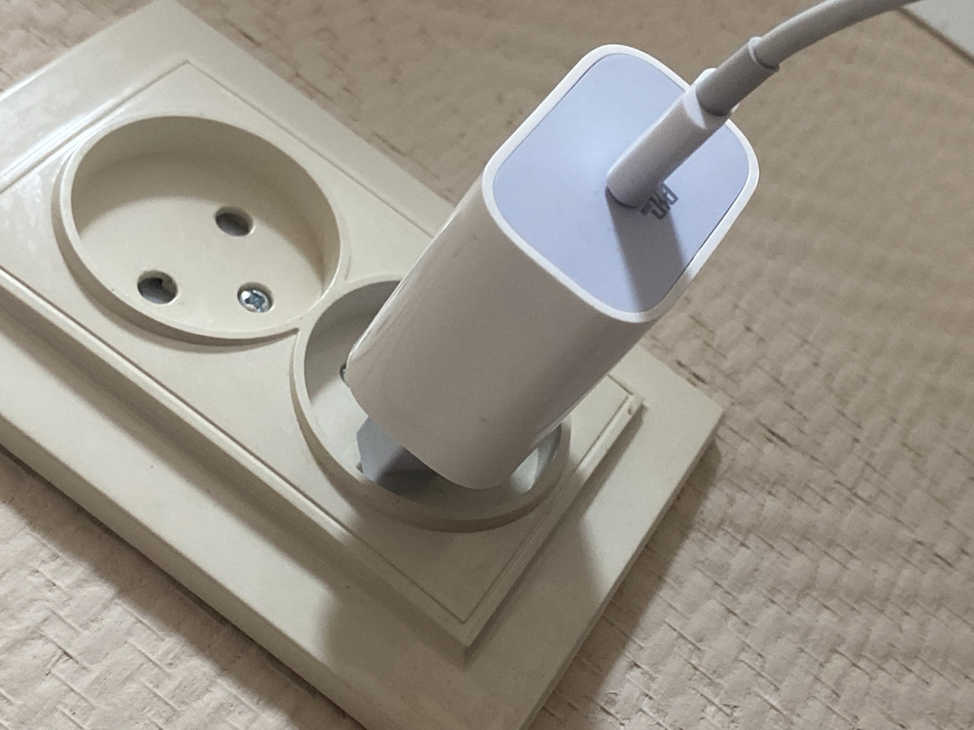 The image shows a close-up, high-angle shot of a power adapter plugged into a wall socket. The socket is a light beige color and has two round holes for prongs and a small screw in the center. The power adapter is also light beige with a light blue accent on the end where the cable connects. The cable is gray and snakes away from the adapter. The wall surrounding the socket is textured, resembling wallpaper or painted plaster, with a light brown or beige color. The lighting suggests an indoor environment. The image does not provide enough information to determine its location in the city of Афанасьево, Russia.