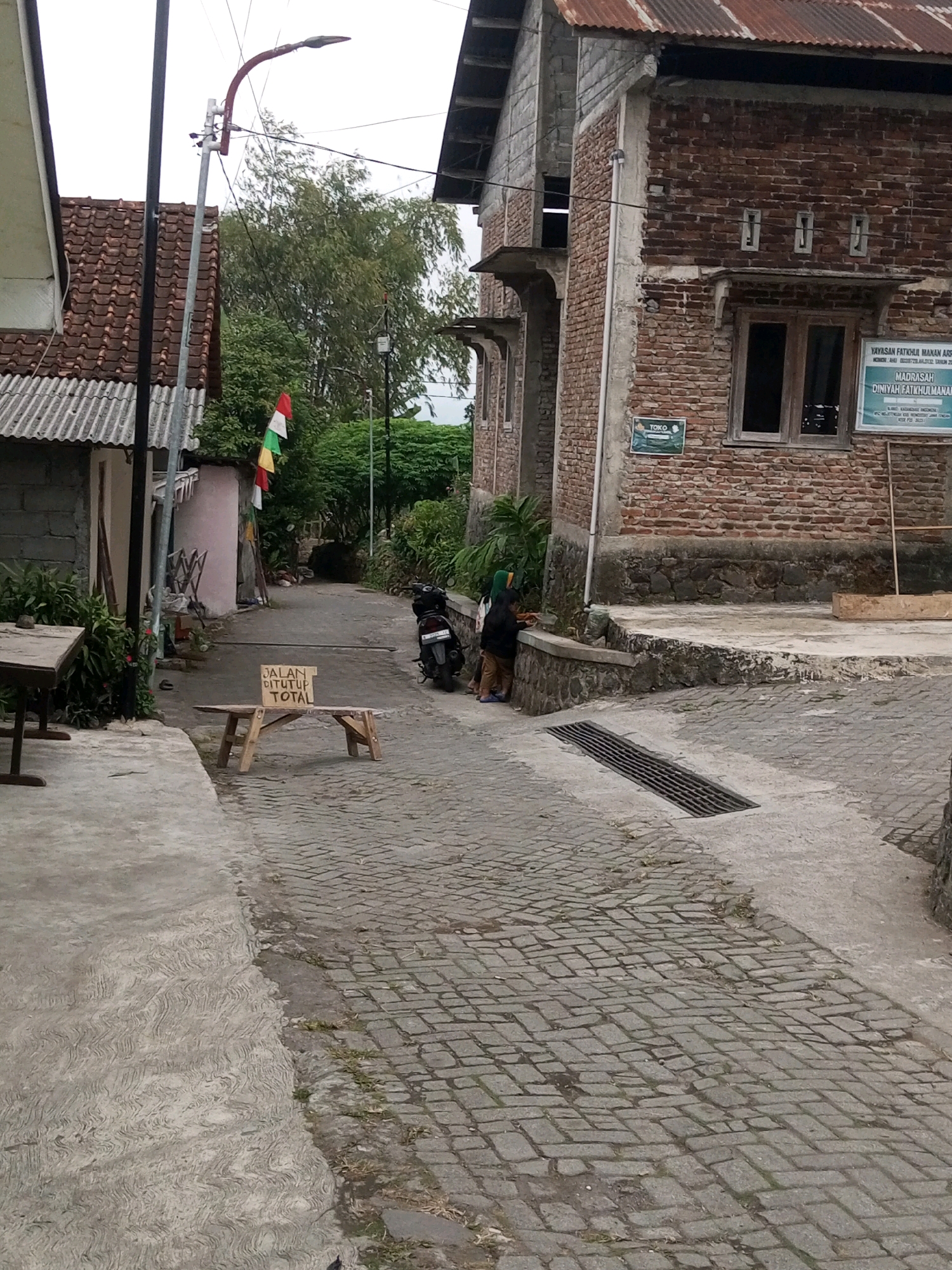 The image shows a narrow, paved street in Andongsili, Indonesia, with buildings on either side. The street is lined with cobblestones and has a concrete sidewalk. On the left, there is a building with a red tile roof and a white corrugated metal wall. A street lamp is visible on the left side of the street. In the middle of the street, there is a wooden sign on a wooden bench that reads "JALAN DITUTUP TOTAL," which translates to "ROAD CLOSED TOTAL." A motorcycle is parked on the right side of the street, and two people, one wearing a green headscarf, are standing near the motorcycle. A brick building with a rusty metal roof is on the right side of the image, with windows and a sign that reads "YAYASAN FATKHUL MA'AN ARSO." The sky is overcast, suggesting it might be a cloudy day.