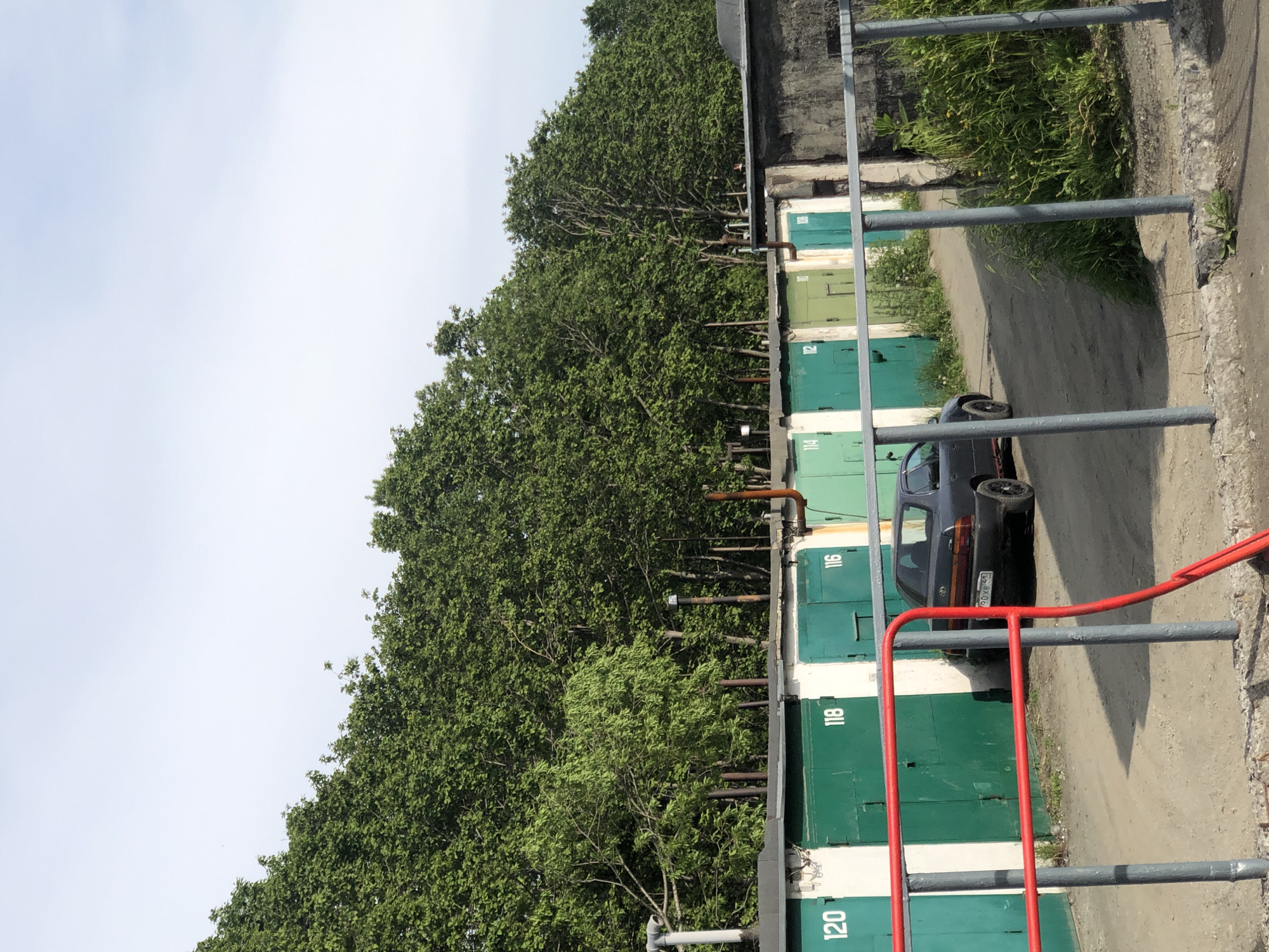 The image captures a row of garages in a residential area of Petropavlovsk-Kamchatsky, Russia. The garages are painted in various shades of green and white, with numbers indicating their respective units, such as 120, 118, and 116. A dark-colored sedan is parked on the dirt road in front of the garages. The background is dominated by a dense line of lush green trees, suggesting a summer or late spring setting. The sky is overcast with a pale, hazy appearance. The scene is framed by a grey metal fence with red railings in the foreground, partially obscuring the view of the garages and the car. The overall impression is one of a typical urban, albeit somewhat rustic, environment in a northern climate.