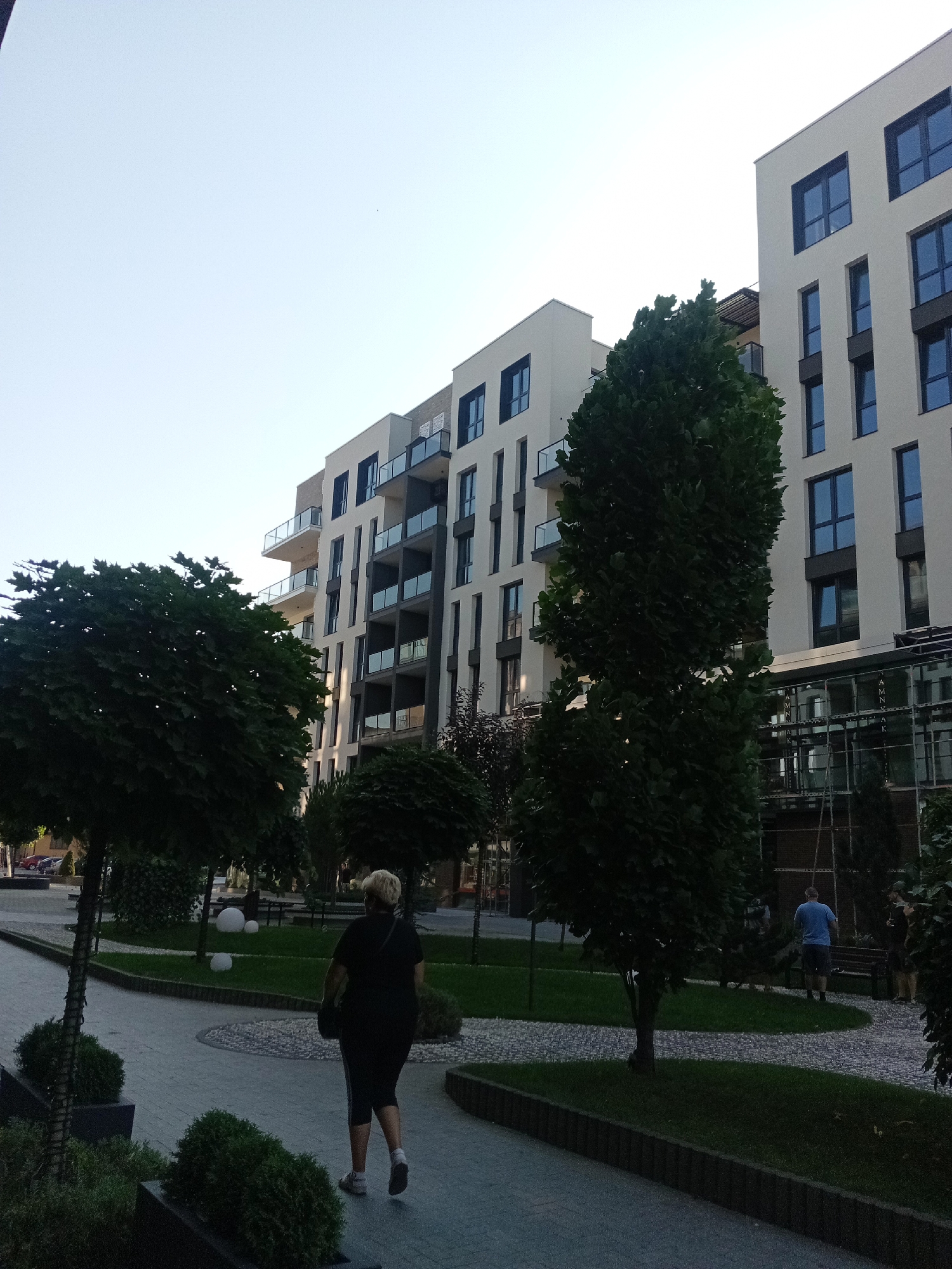 A medium shot captures a modern apartment complex with a clear sky overhead. The building's facade is light-colored, punctuated by dark-framed windows and balconies with glass railings. In the foreground, a woman with blonde hair, wearing a black top and pants, walks away from the camera on a paved walkway. She carries a dark bag over her shoulder. To her left, a tall, slender tree stands, its leaves obscuring part of the building. Several smaller trees and manicured bushes are scattered throughout the scene, some surrounding decorative white orbs on a patch of grass. A man in a blue t-shirt and shorts is visible in the background, seated on a bench near another tree. The environment suggests a well-maintained urban residential area.