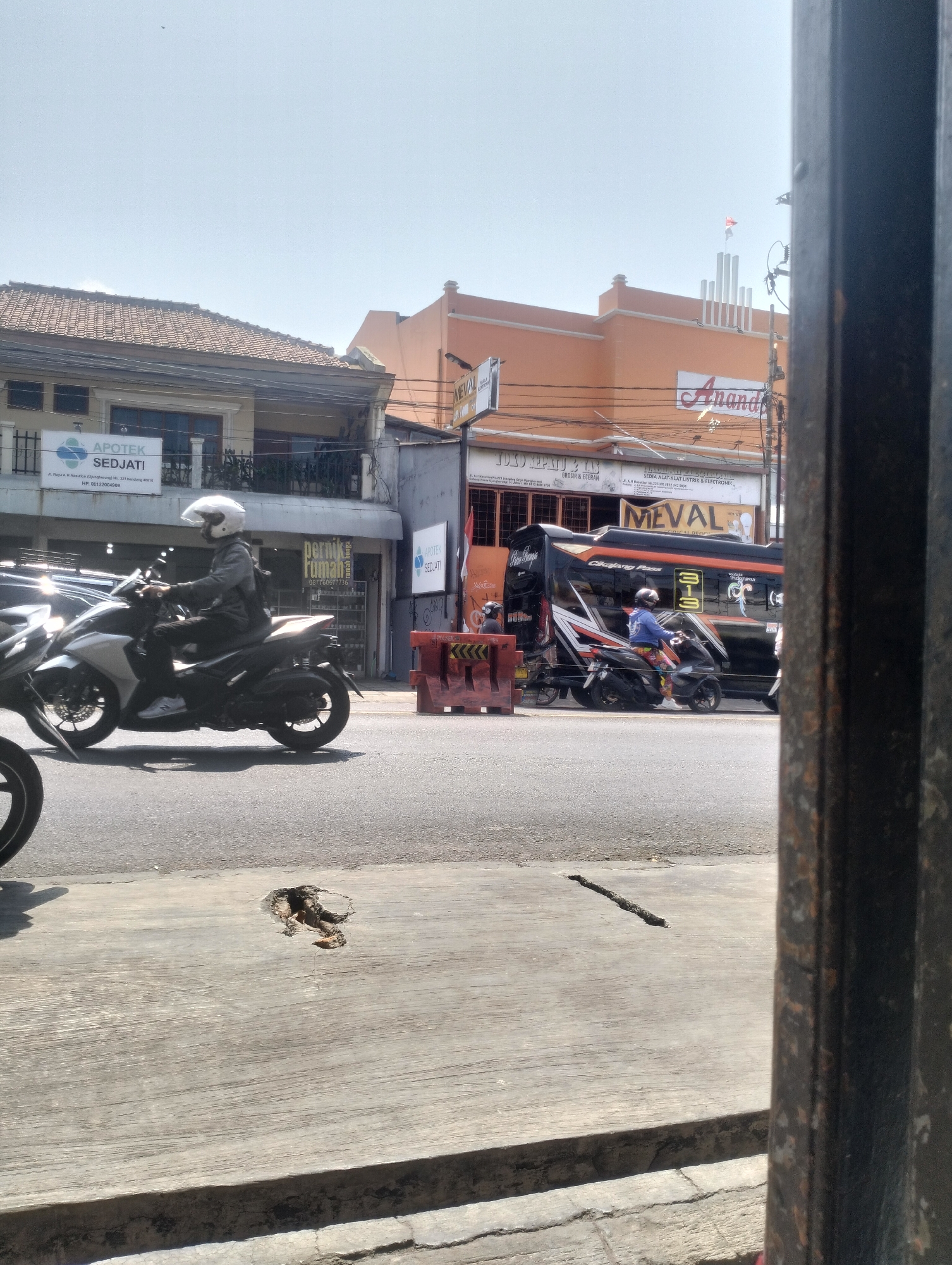 A street scene in Kel. Pasir Jati, Indonesia, shows a bright, sunny day with clear skies. The foreground features a concrete sidewalk with a large crack and a smaller fissure. Beyond the sidewalk, a paved road runs horizontally, with two motorcycles and a bus visible. The first motorcycle, a white model, is ridden by a person wearing a helmet and dark clothing, with a backpack. The second motorcycle, black, is also ridden by a person in a helmet and colorful pants. The bus, black with orange and white accents, has "MEVAL" written on its side and appears to be a passenger bus. Several storefronts line the street, including "APOTEK SEDJATI" (pharmacy) and a business with the sign "PERNIK RUMAH" (home furnishings). Another building with an orange facade has a sign reading "Ananda" in red script. The environment suggests a commercial area within a city.
