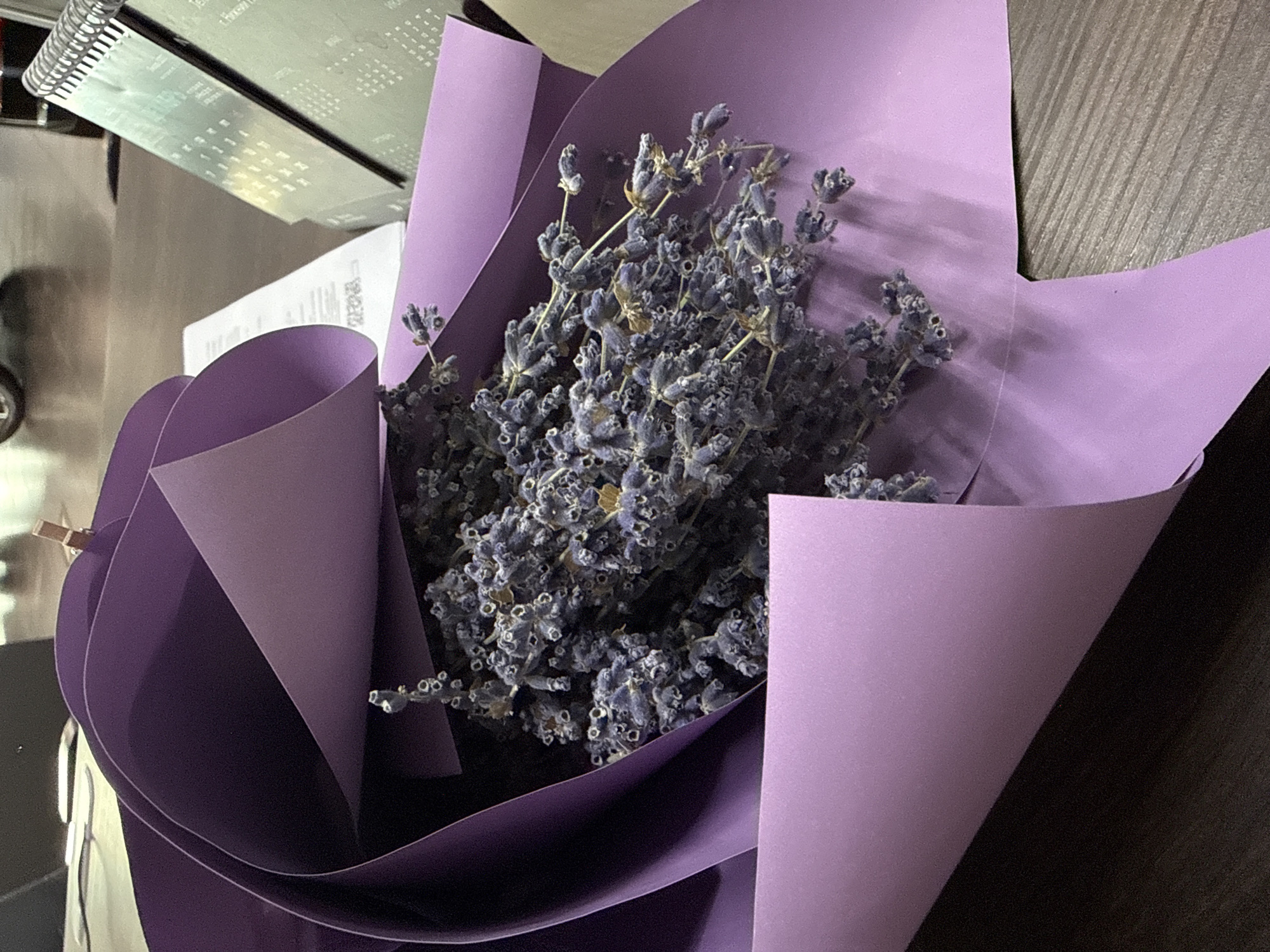 A bouquet of dried lavender is partially wrapped in purple paper, resting on a dark wooden surface. A desk calendar is visible in the background, along with what appears to be a document or receipt. The scene is indoors, likely an office or home setting, in Nizhny Novgorod, Russia.