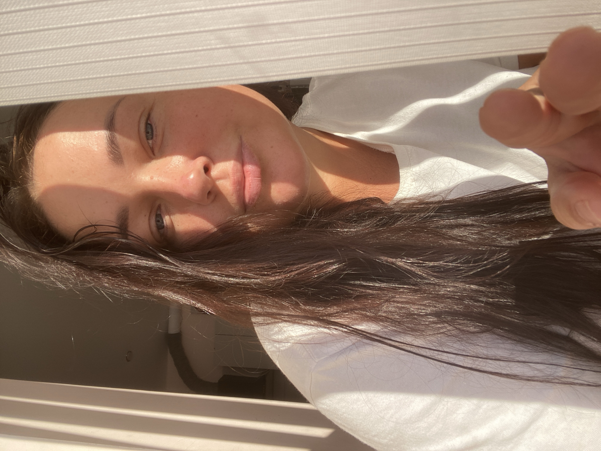 A woman with dark hair is looking at the camera. She is wearing a white t-shirt. Sunlight is casting shadows across her face, highlighting her features. She is reaching out towards the camera with her right hand. The background shows vertical blinds and a hint of a ceiling fan. The lighting suggests the image was taken during the day, possibly indoors near a window.