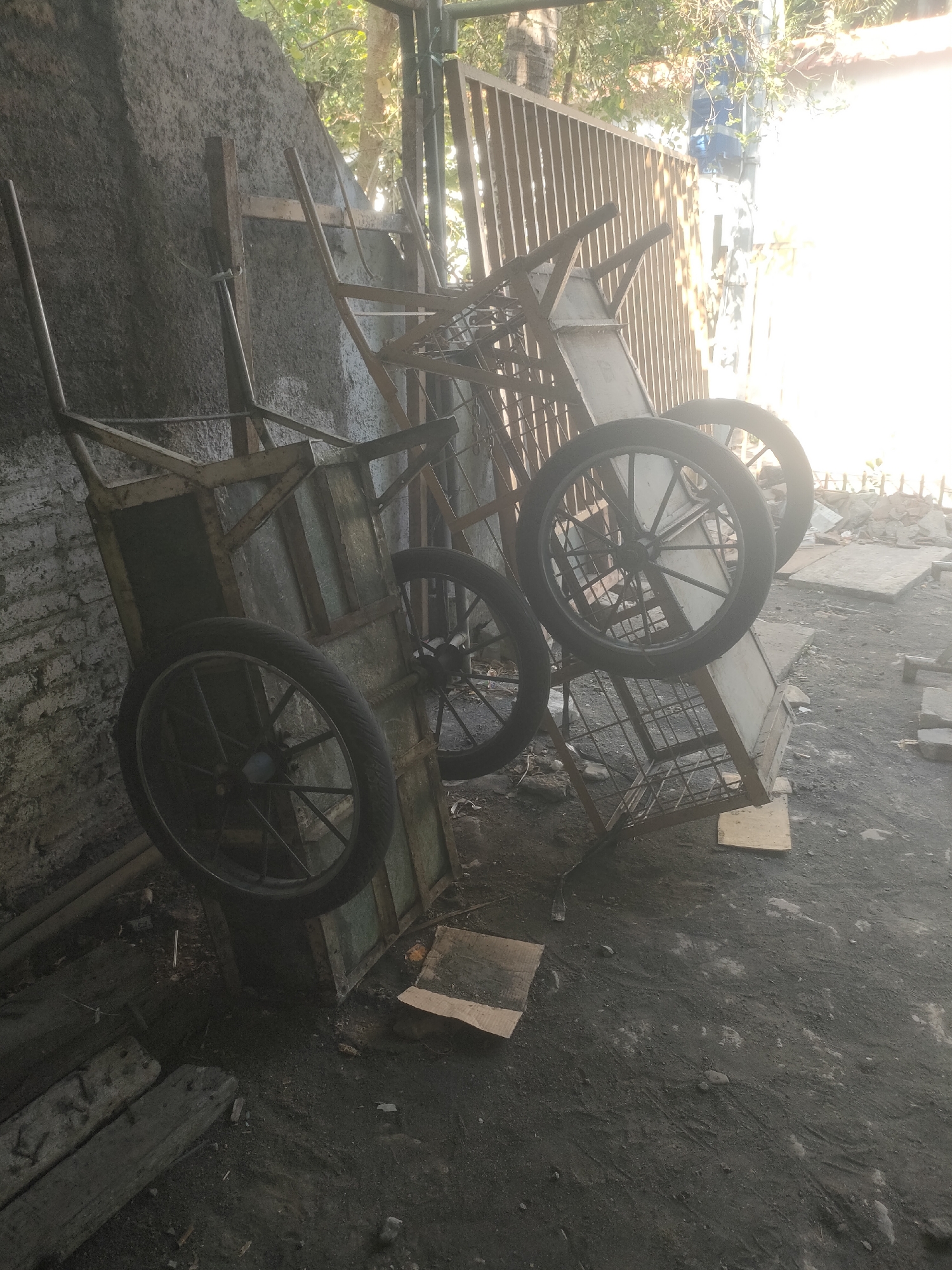 This image depicts an outdoor scene, likely a workshop or storage area, in Surabaya, Indonesia. The foreground is dominated by two old, utilitarian wheelbarrows. The wheelbarrow on the left is upright and appears to be made of metal with wooden planks forming its cargo bed. It has one large, spoked wheel with a rubber tire. The second wheelbarrow, positioned behind and to the right of the first, is tilted on its side. It also has a spoked wheel with a rubber tire, and its cargo bed seems to be a metal grate.
In the background, a rustic, vertical slatted fence or gate is visible, with sunlight streaming through it. To the left of the wheelbarrows, a rough, concrete or stucco wall, possibly part of a building, rises up. The ground is covered in dark, dusty earth, with scattered debris including pieces of wood and what appear to be broken concrete blocks. The overall atmosphere is one of disuse and a slightly neglected industrial or construction environment. The natural light suggests it is daytime. There are no people visible in the image.