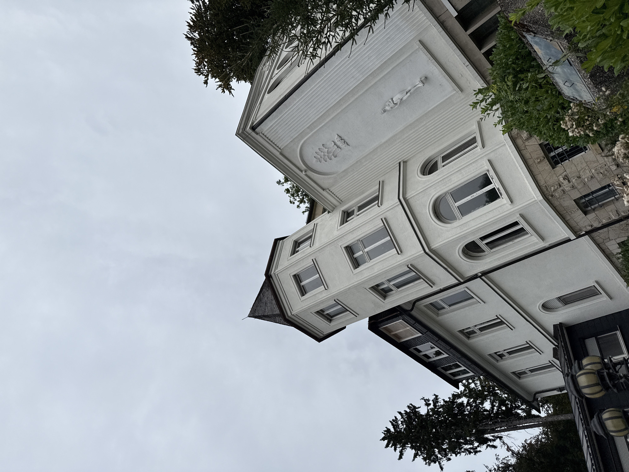 A low-angle shot captures a white, multi-story building with a pointed dark roof and a turret, set against an overcast sky. The building features numerous windows, some arched. Decorative relief sculptures adorn the facade. To the left of the building, a tall, dark evergreen tree is visible. In the foreground, lush green foliage and a stone-clad section of the building are present. Two ornate lampposts with amber glass globes stand on the left side of the image. The overall scene suggests a residential or institutional building, possibly in a town setting given the hints of a street or pathway in the foreground. The location is identified as Bad Sachsa, Germany.