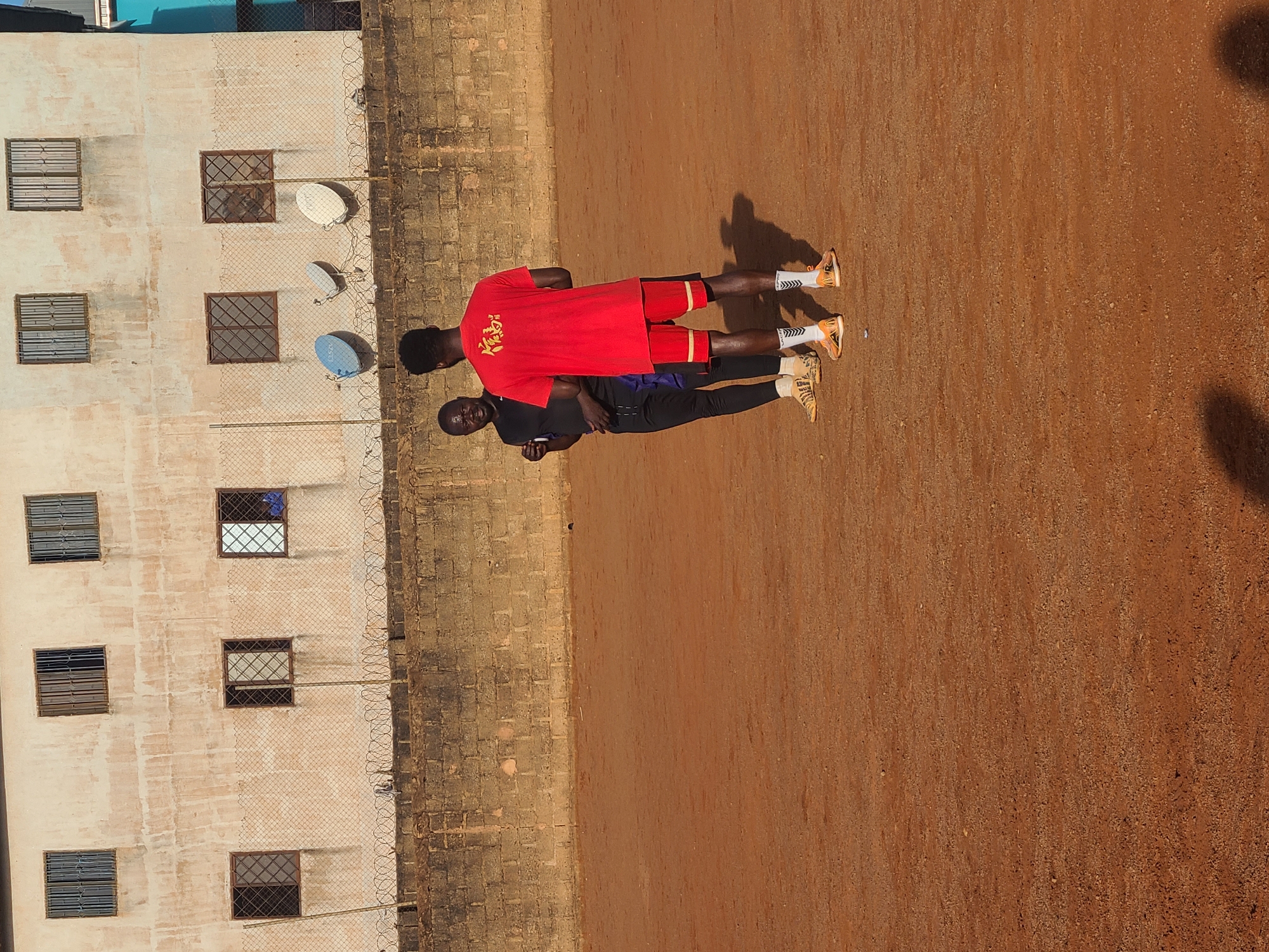 The image displays two men standing outdoors on a reddish-brown, dusty or dirt ground under bright sunlight. The man in the foreground is seen from behind, wearing a red short-sleeved t-shirt with a gold-colored design on the back, and matching red shorts with white stripes. He wears orange and white athletic shoes with patterned white socks. The second man stands slightly behind and to the left of the first, wearing a dark-colored t-shirt and long pants with light-colored athletic shoes. His face is visible, looking towards the viewer. Both men have dark skin and short, dark hair.
Strong shadows of the men are cast on the ground, indicating clear weather and strong sun, suggesting either morning or late afternoon.
To the left of the men, a multi-story building with a light-colored, possibly stucco, facade is visible. It features several rectangular windows, each secured with metal grates. A chain-link fence, topped with barbed wire, runs parallel to the building. Three white satellite dishes are mounted on the building wall, seen through the fence; the letters "CSP" are visible on one of them. The setting appears to be an urban or semi-urban environment, possibly a residential or commercial area. There are no clear emotional expressions or specific activities other than standing.