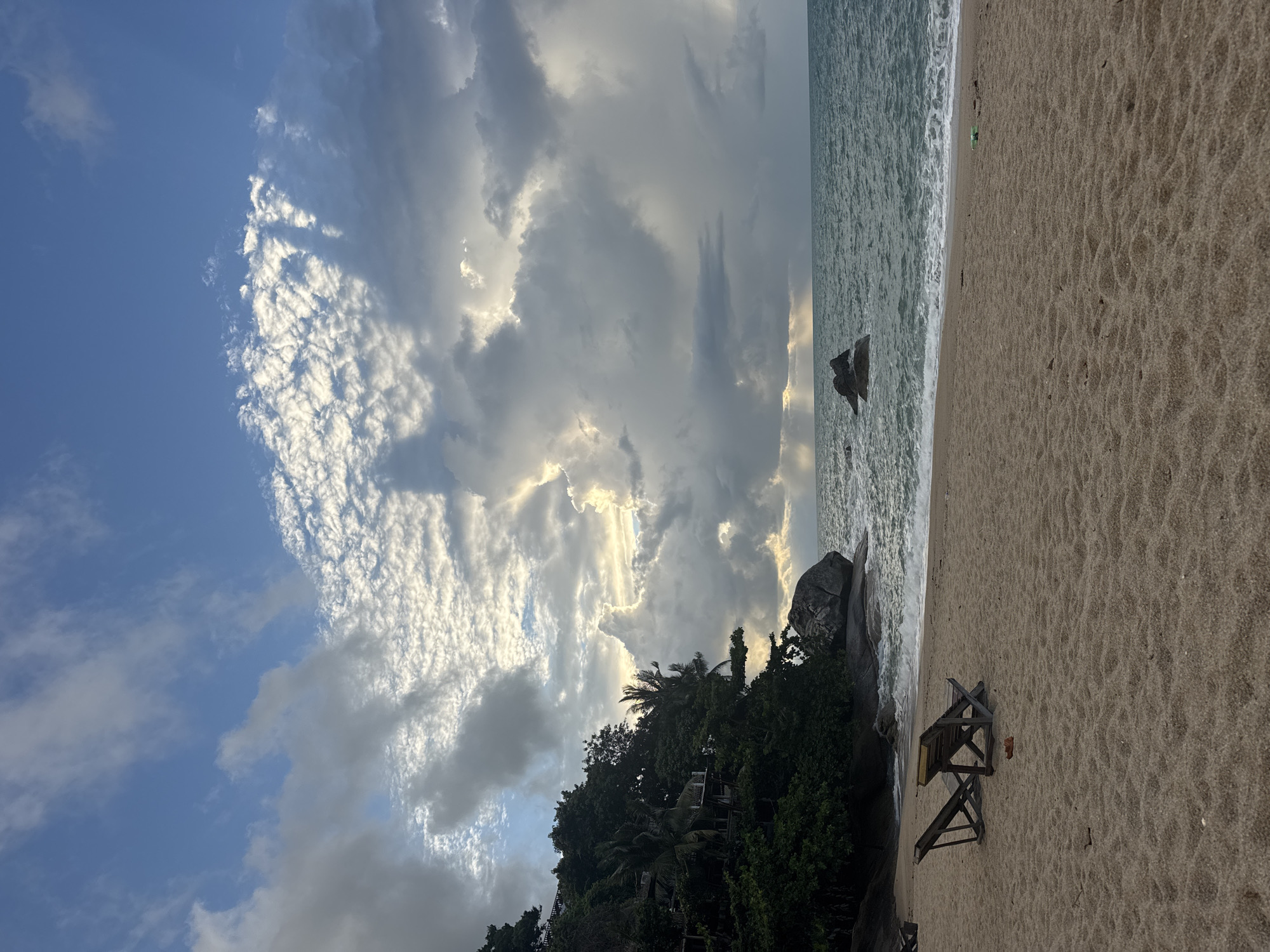 The image shows a beach scene in Baan Thong Nang, Thailand. The beach is sandy, with a weathered wooden beach chair visible on the right side. Vegetation, including trees and possibly buildings, is situated behind the beach in the lower-center. In the background, the ocean meets the shoreline, with waves. Two rocks protrude from the ocean. The sky is filled with a mix of white and gray clouds, some with a patterned appearance, and sunlight is filtering through the clouds.