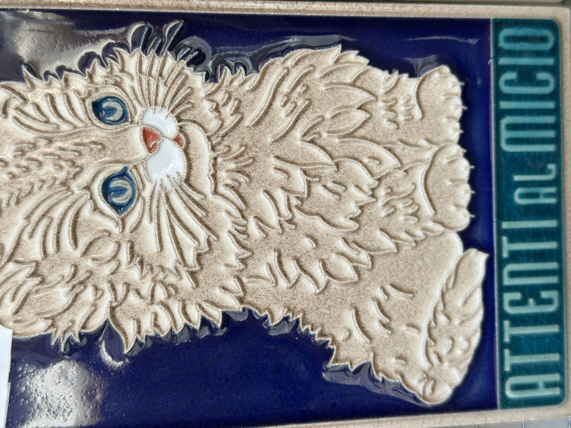 A detailed close-up photograph showcases a ceramic tile featuring a cat design, accompanied by Italian text, likely found in Maiori, Italy.
The tile consists of a dark blue, glossy background. At the center of the tile is a beige cat relief with long fur, a white muzzle with a small, red nose, and piercing blue eyes. To the right of the cat, text appears vertically in blue and light green blocks that reads "Attenti Al Micio."
