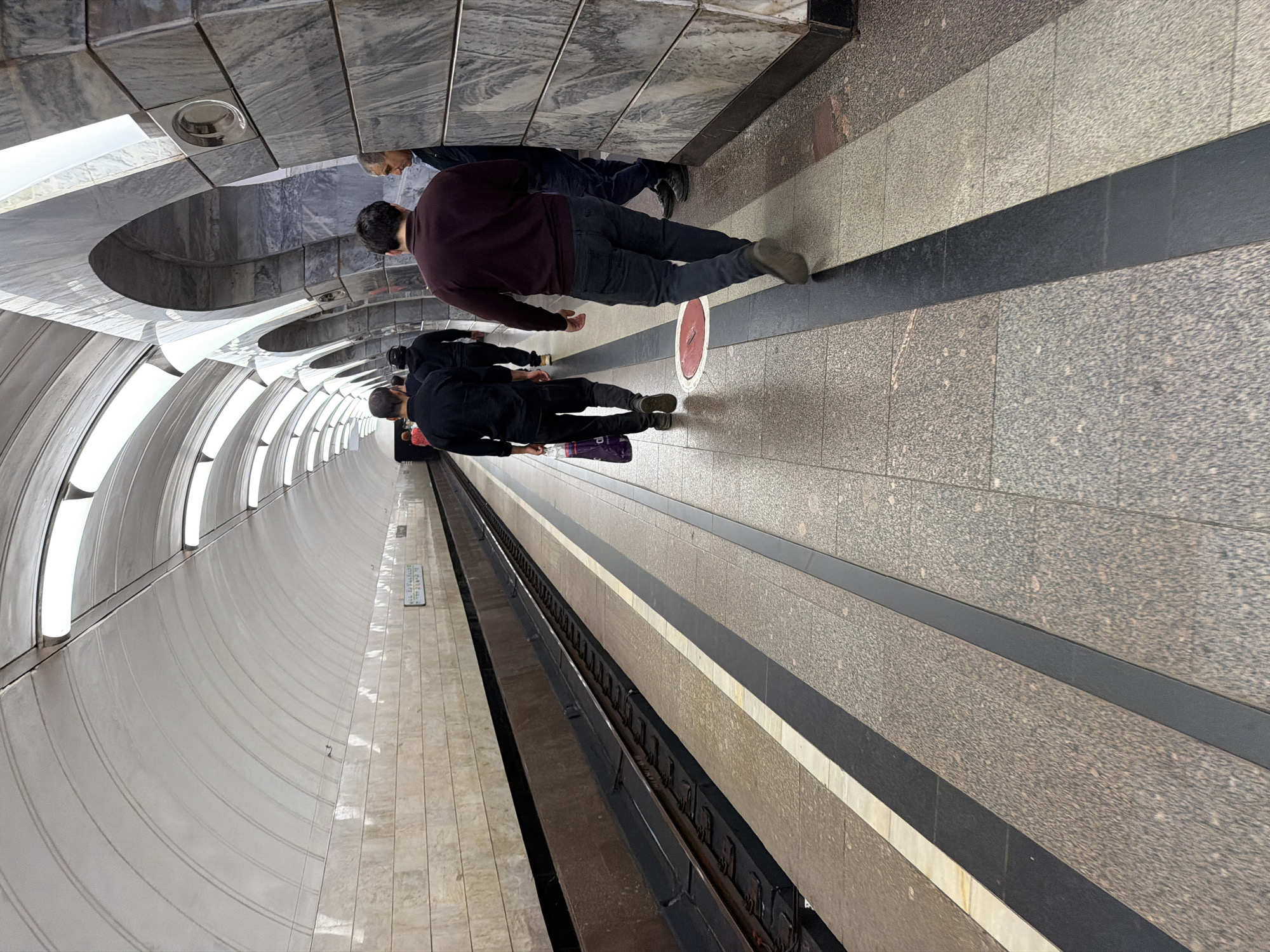 This is a medium-angle shot of a subway platform in Moscow, Russia. The platform is made of granite tiles with black and white stripes marking the edge. The walls are lined with beige tiles, and the ceiling is a white, arched structure with integrated lighting. In the background, the subway tracks are visible.
Several people are walking away from the camera along the platform. The person in the foreground on the right is wearing a dark maroon sweater and dark jeans. To their left, a man in a black jacket and dark pants is walking. Further down the platform, a man in a black hoodie and dark pants is visible. To the left of the man in the hoodie, another person in a black shirt and pants is carrying a purple shopping bag. Two more individuals are further down the platform, one in a black jacket and the other in a dark shirt. On the far right, an older man with short gray hair is standing near a marble-clad pillar. The overall scene suggests a typical day at a Moscow subway station.