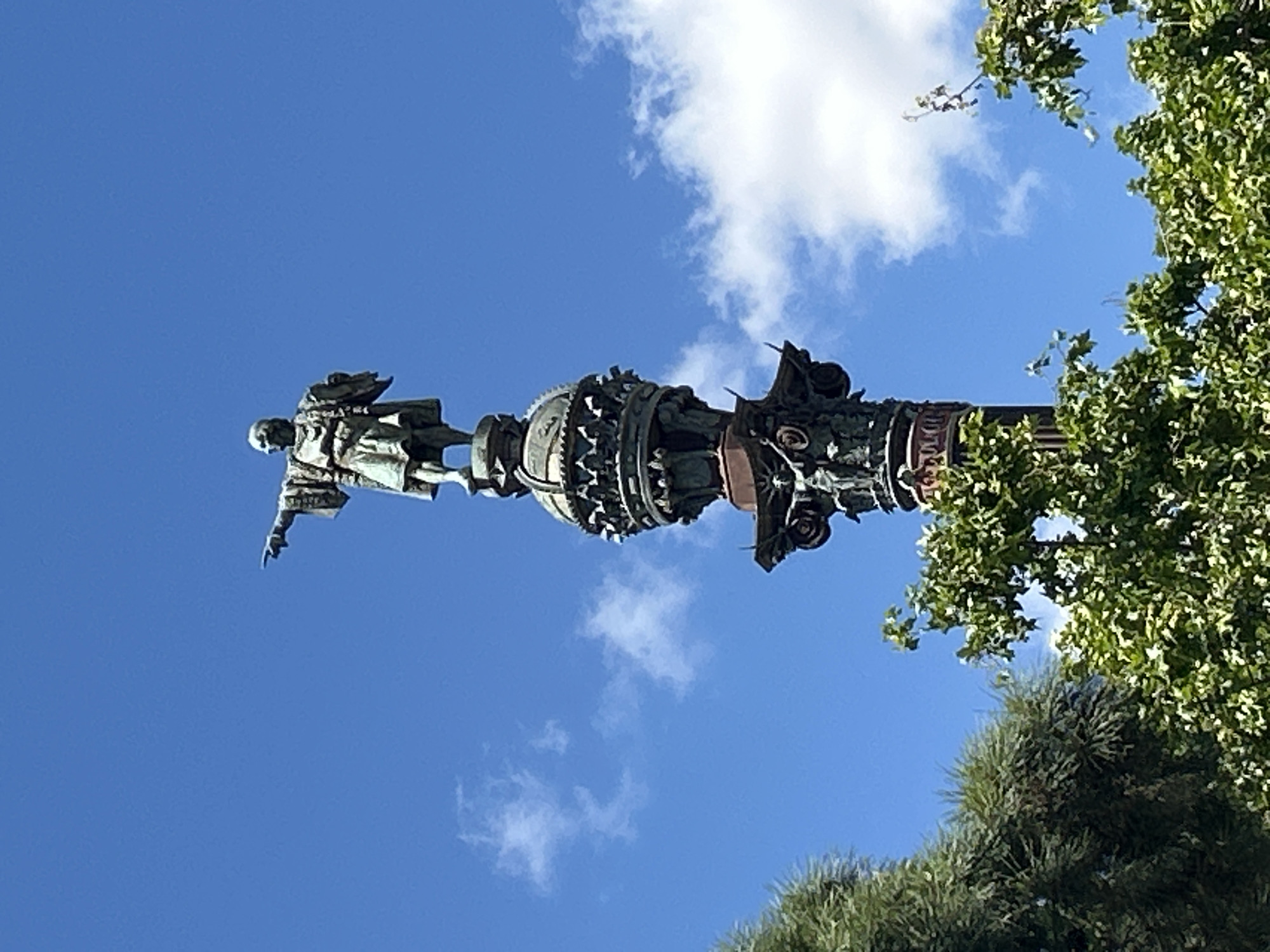 The image shows the Columbus Monument in Barcelona, Spain, viewed from a low angle. The bronze statue of Christopher Columbus atop a decorated column is prominently visible against a blue sky with scattered white clouds. The statue faces left, appearing to point with its right hand. The column itself is ornate and detailed. Green trees and foliage fill the right side of the frame, partially obscuring the column.