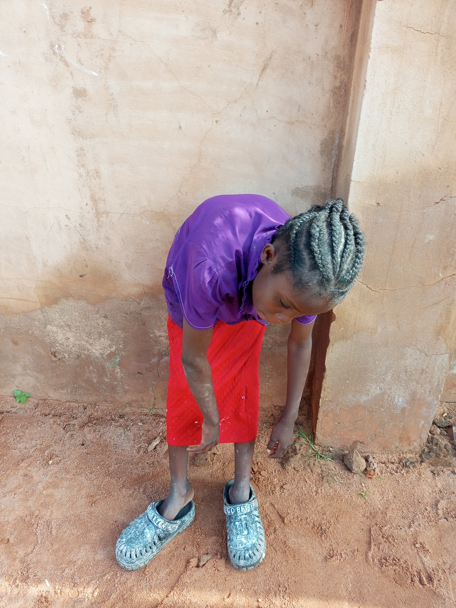 A young Black girl with braided hair, wearing a purple shirt and a red skirt, stands on sandy ground in front of a light brown, cracked stucco wall in Tempure, Nigeria. She is bent over, looking down at her feet, which are clad in grey, patterned clogs. One clog has the words "GO BETTER" visible. A small green plant sprouts from the ground to the left of the girl. The lighting suggests it is daytime, with harsh shadows cast by the girl and the wall.