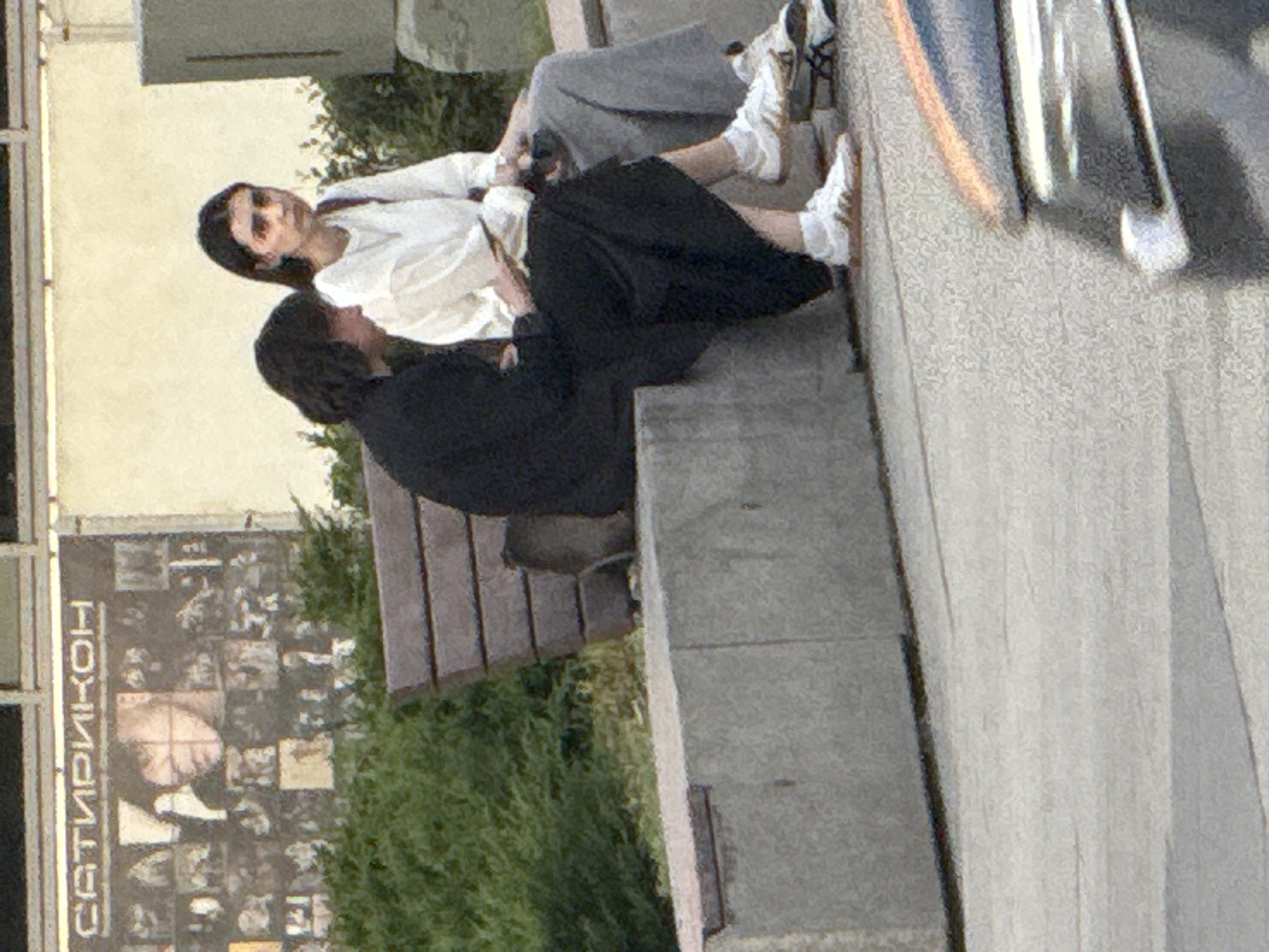 Two people are sitting on a bench outside in Moscow, Russia. One is wearing a white shirt and gray pants, and the other is wearing a black shirt and black pants. Both have on white sneakers. A poster is to the left of them. The pavement is concrete.
