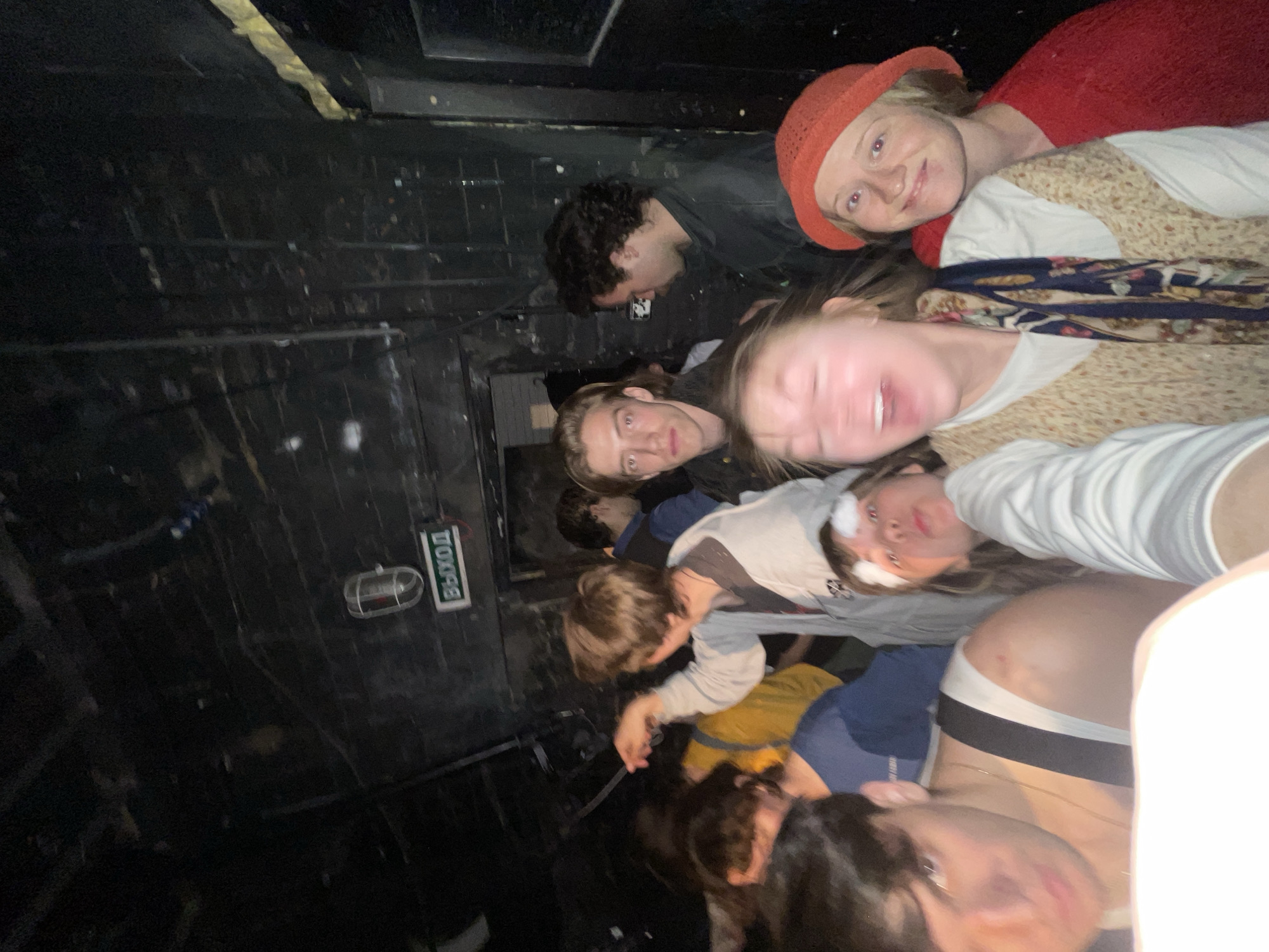 A group of people are taking a selfie in a dark, enclosed space. The space appears to be a narrow corridor or small room with a dark, possibly metallic, wall. A sign with the word "ВЫХОД" (EXIT) in Cyrillic script is visible on the wall, along with a small light fixture.
The people are clustered together, looking into the camera. They have a variety of clothing on.
Based on the presence of the Cyrillic sign, the location is likely in a Russian-speaking country, potentially in the city of Moscow, Russia.