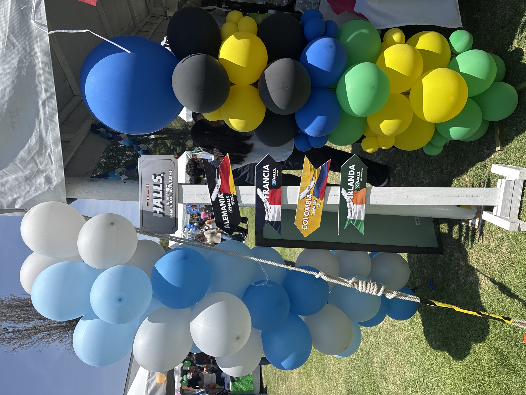 This media file captures an outdoor event in San Andrés Cholula, Mexico, on a bright and sunny day. The scene is dominated by vibrant balloon decorations and promotional displays. Large clusters of balloons in various colors—white, light blue, dark blue, black, yellow, and green—are arranged around white tent structures, suggesting a festive atmosphere.
Prominently featured are several pennant flags representing different countries, including "ALEMANIA" (Germany), "FRANCIA" (France), "COLOMBIA," and "IRLANDA" (Ireland). Each flag is associated with the "HALLS" brand and a specific flavor: "HALLS MENTA" (Mint) for France, "HALLS LIMON" (Lemon) for Colombia, and "HALLS YERBABUENA" (Spearmint) for Ireland. A large sign for "HALLS" also displays the slogan "UN RESPIRO, UN MUNDO DE AVENTURAS" (A breath, a world of adventures).
Several people are visible in the background, moving around or standing near other stalls, suggesting a public gathering, fair, or promotional event. The ground is covered in green grass, and the strong sunlight casts clear shadows. A thick rope with a segment of black and yellow stripes is visible in the foreground, possibly part of an activity area or a decorative boundary. The overall impression is that of a lively outdoor event, likely a cultural festival or a marketing campaign for Halls candies, focusing on international themes and flavors.