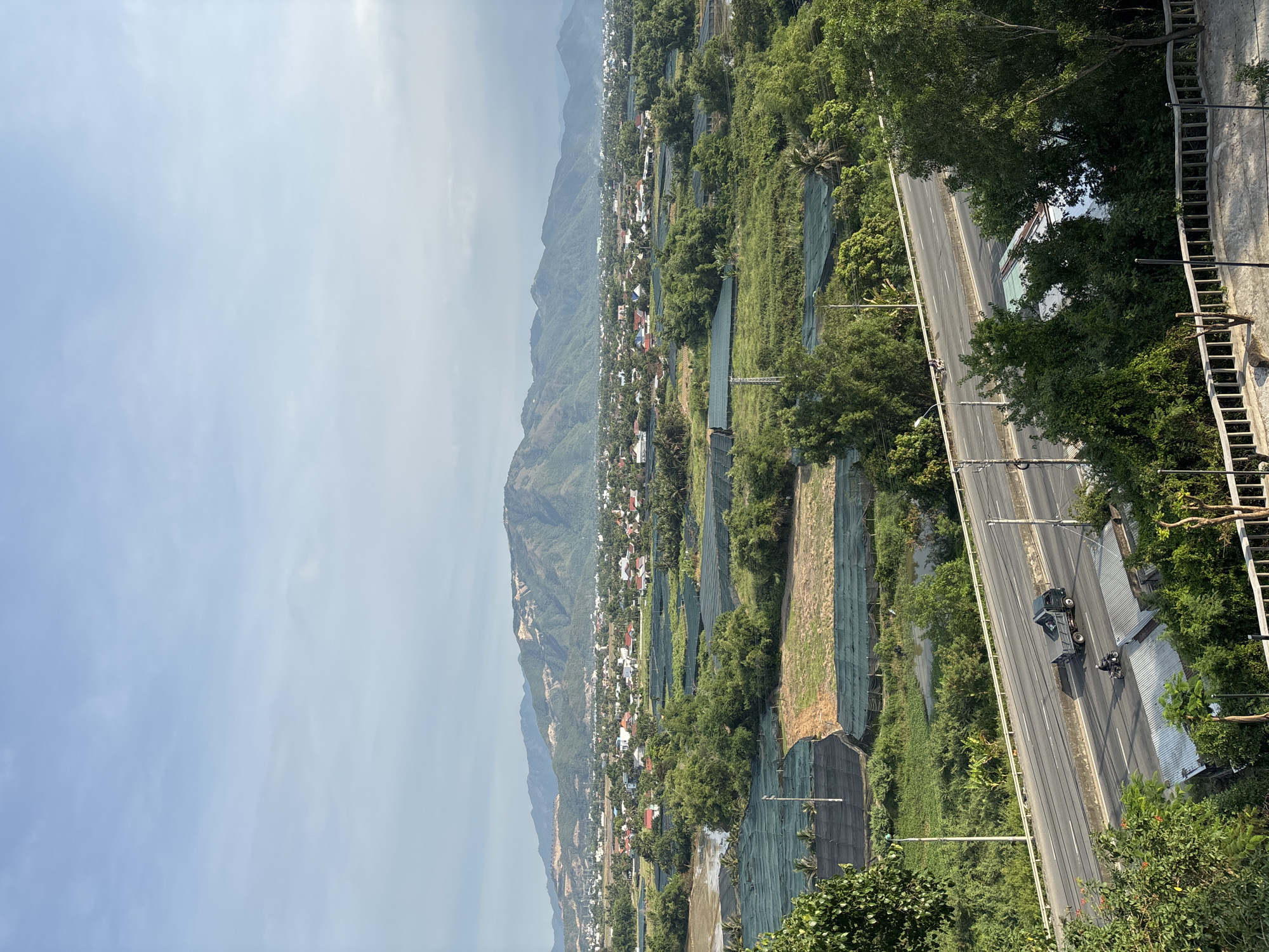This is a high-angle, eye-level, full shot of a landscape in Tay Nha Trang Ward, Vietnam, featuring a town nestled between rolling green fields and mountains under a clear sky. In the foreground, a road runs horizontally across the frame, with a dark grey truck and a motorcycle traveling on it. The road is bordered by lush green trees and vegetation. Beyond the road, the landscape transitions into agricultural fields, many of which are covered by green netting structures. A body of water, possibly a pond or irrigation channel, is visible amidst the fields. In the midground, a town with numerous houses, many with red roofs, is spread out. The background is dominated by a range of mountains with sparse vegetation and visible rock formations. The sky is a pale blue with scattered clouds.