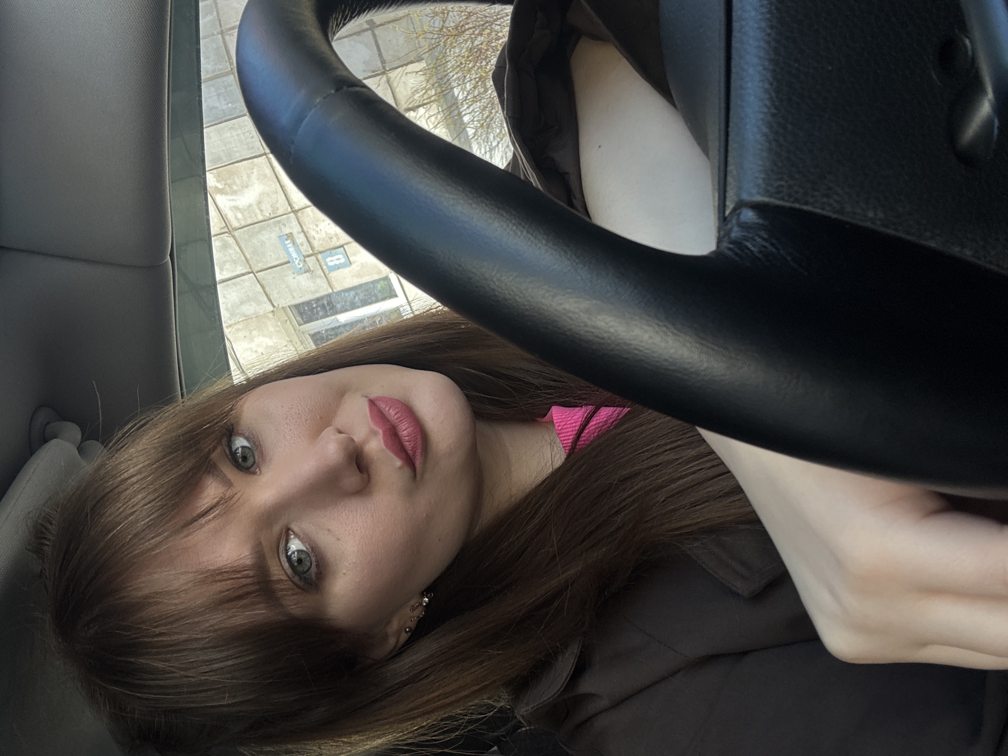The image displays a young woman inside a car, looking directly towards the camera. Her head is tilted, and she has long, straight brown hair with bangs that frame her forehead. Her eyes are a light blue-green, and she is wearing pink lipstick, with a subtle, neutral expression. A bright pink collar or top is visible around her neck, worn beneath a dark jacket. A delicate earring with small dangling stones adorns her left ear.
A black steering wheel is prominent in the foreground, partially obscuring the upper right side of the image and her torso. Through the car window, bright daylight illuminates an outdoor scene. The ground outside appears to be paved, resembling a sidewalk or parking lot, with visible square paving stones. Bare tree branches are visible against the sky, suggesting a colder season.
On the paved ground, a blue rectangular marking with white text is discernible. Part of the text clearly reads "ПАРКОВКА" (Russian for "PARKING"), and a white number "8" or infinity symbol is also visible within the blue marking. The setting appears to be an urban or suburban environment, consistent with the provided location hint of Chaikovsky, Russia. The bright lighting indicates it is daytime.