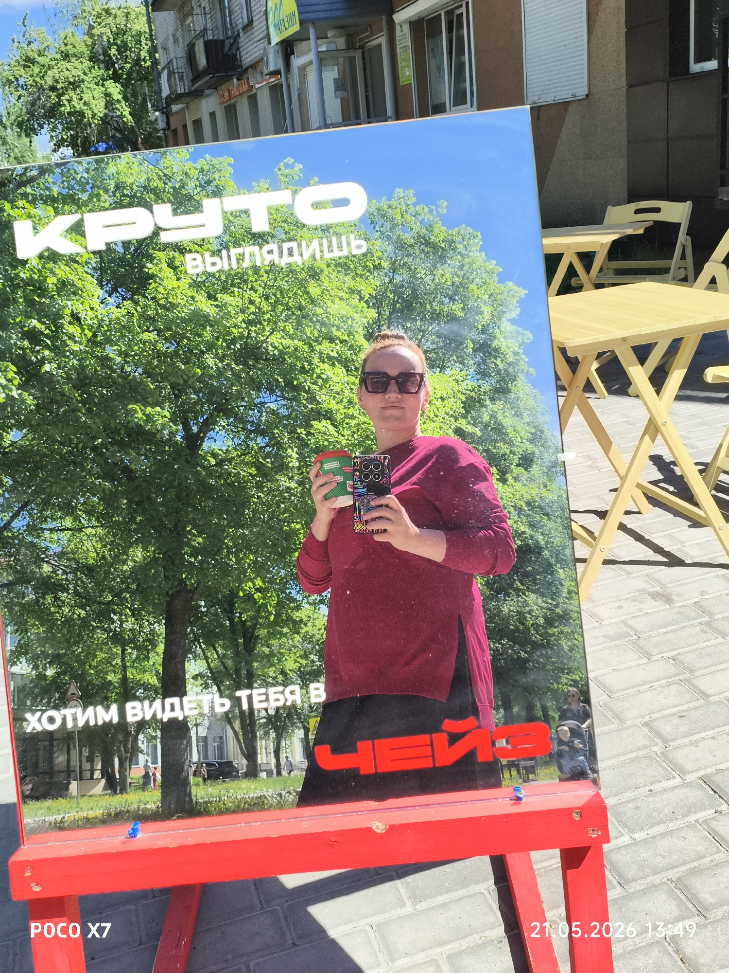 The image shows a woman reflecting in a mirror outside a building. The mirror is propped on a red stand with Russian text, "КРУТО выглядишь Хотим видеть тебя в…". The reflection shows the woman holding a cup and a phone. The background reflection shows trees, a street, and buildings. To the right of the mirror are yellow tables and chairs. The photo includes a timestamp and camera information at the bottom. The location appears to be an urban environment, possibly a cafe or restaurant exterior in Unknown, Unknown.
