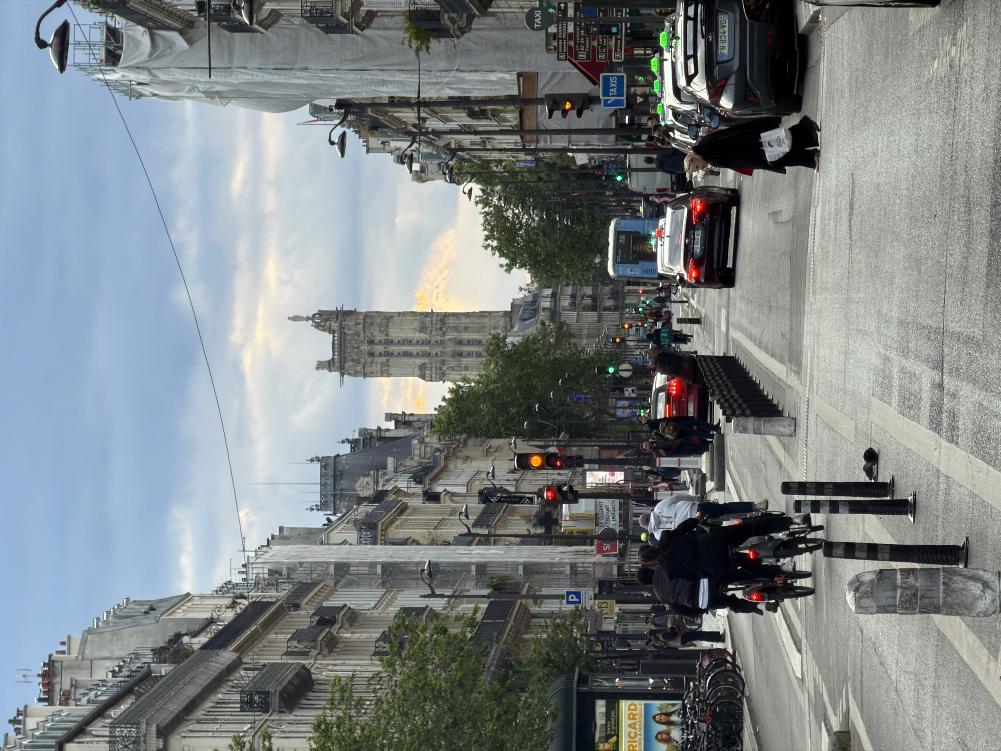 The image shows a street scene in Paris, France. A tall, ornate stone building dominates the view, framed by lower buildings lining both sides of the street. The sky is light blue with some clouds. Traffic is present on the street, including cars, buses, and motorbikes with multiple riders. Pedestrians are visible on the sidewalks. Roadside features include traffic lights, street signs (including "TAXI" signs), parking signs, striped bollards, and bicycle racks. Advertisements, including a large one for RICARD, are also visible. Scaffolding covered in white material is present on some buildings. A woman in dark clothes is crossing the street while holding some printed materials.