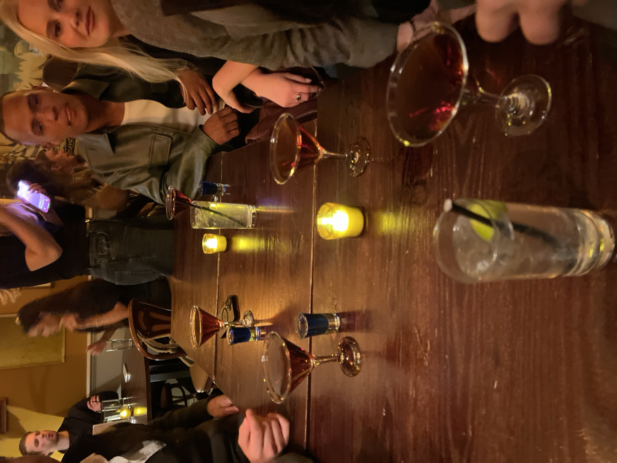 A group of people are sitting at a dark wooden table, illuminated by candles. There are several cocktails in martini glasses and highball glasses on the table, along with a couple of small liquor bottles. Two individuals are closely positioned at the top left of the frame, while others are partially visible around the table. A person is looking at their phone towards the lower-left corner of the image. The specific location in London is not evident from the image.