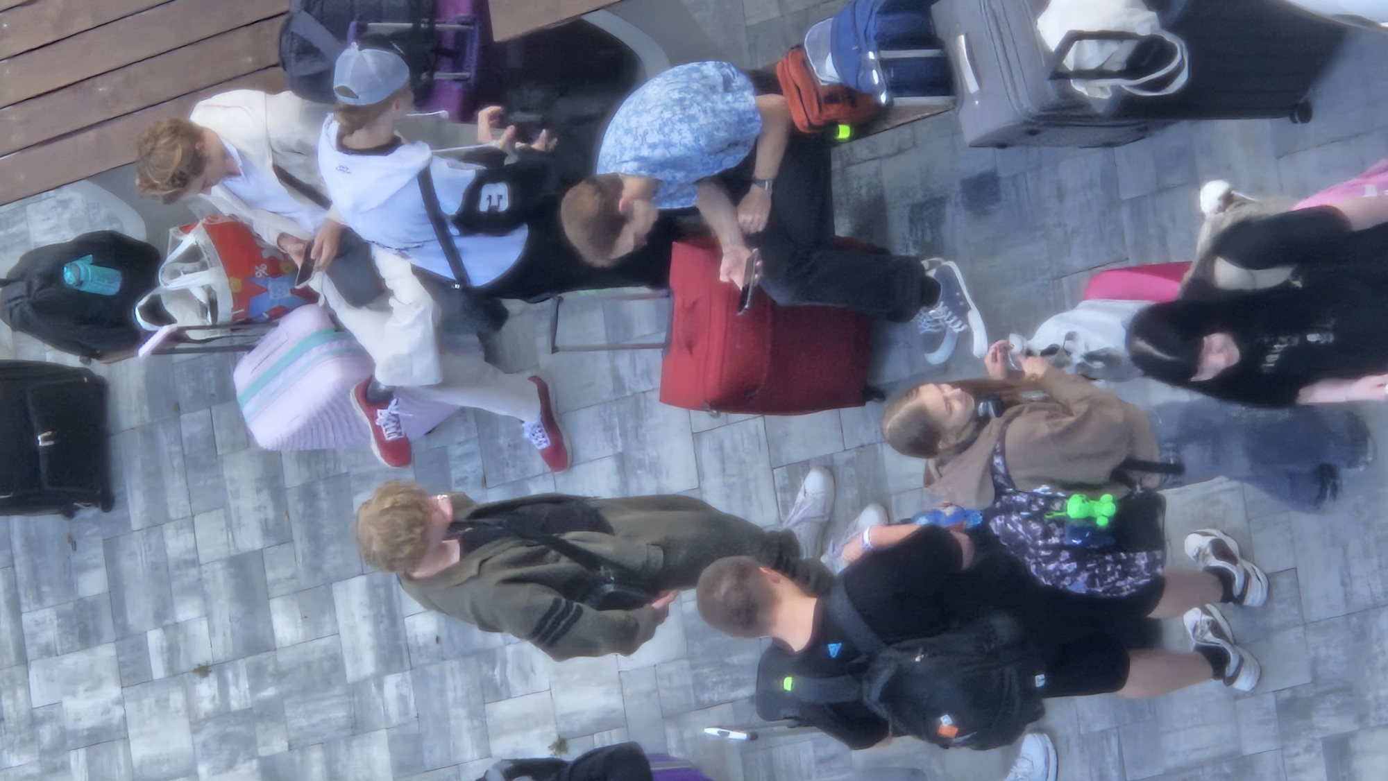 The image depicts a group of young people with luggage, suggesting a travel or departure scenario. The setting appears to be an outdoor paved area, possibly a train station or an airport waiting area, given the presence of various suitcases and backpacks. The ground is paved with rectangular grey and white tiles arranged in a grid pattern.
In the foreground, a person with short, light brown hair wearing a green jacket and dark pants is standing and looking downwards, possibly at their phone or an object in their hand. Next to them, a person with short blonde hair is seated on a red suitcase and looking at their phone.
Further back, a person in a white jacket and beige pants is seated, also engrossed in their phone. To their right, another person wearing a white and blue patterned shirt is seated with a phone in their hand.
In the middle ground, a person with brown hair and a brown hoodie is standing and interacting with their phone. They are wearing jeans and carrying a backpack adorned with a bright green charm. To their right, a person with long dark hair is visible in the lower right corner of the frame, looking downwards.
Various luggage items are scattered around, including black backpacks, a pink suitcase with teal stripes, a red suitcase, a purple suitcase, and a grey suitcase. Some of the luggage appears to be wheeled. The lighting suggests it is daytime, possibly overcast or late afternoon, as there are no strong shadows. The overall impression is of people waiting or preparing for travel in an urban outdoor environment.
The provided image does not contain any visual cues that would allow for its specific identification to the city of Prešov, Slovakia.