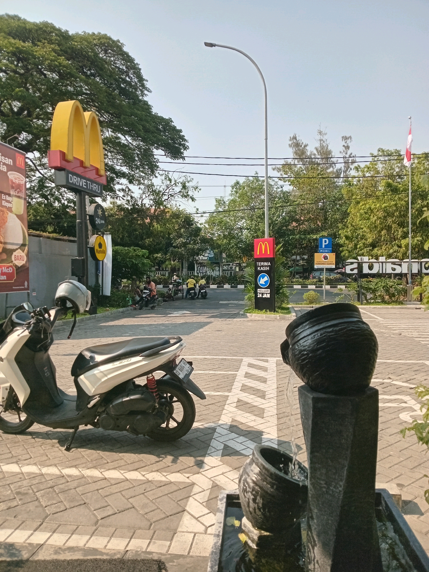 The image captures a view of a McDonald's restaurant with a drive-thru service in Surakarta, Indonesia. In the foreground, a white and black scooter is parked on a patterned brick surface. A helmet rests on the handlebars of the scooter. To the right, a dark gray stone fountain with two decorative pots is visible, with water flowing between them.
In the middle ground, the McDonald's signage is prominent, featuring the iconic golden arches and a "Drive Thru" sign. Several people on motorcycles are present in the background, suggesting a busy day. A street lamp arches over the scene, and power lines are strung across the sky. Trees line the background, and a flag is visible to the right of the frame. The environment appears to be an outdoor public space, likely a commercial area given the presence of the fast-food restaurant.