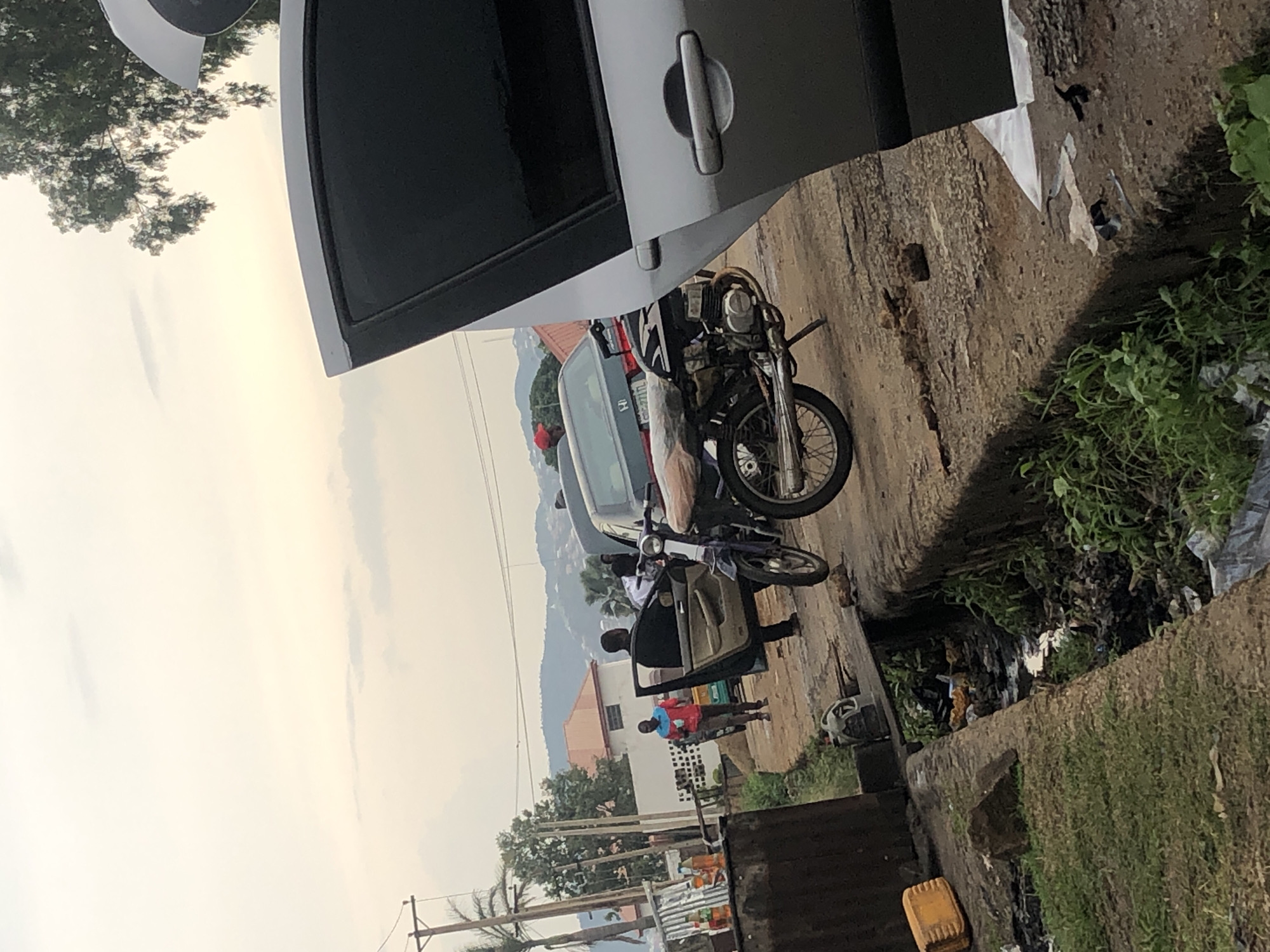 The image depicts a street scene in Jos, Nigeria, under an overcast sky. In the foreground, a concrete drainage ditch filled with debris and stagnant water runs alongside a dirt road. A silver car, partially visible from the side with its passenger door open, dominates the right side of the frame. To the left of the silver car, a motorcycle is parked on the road, with its rear wheel and part of the engine visible.
On the road, a Black man in a black suit is exiting a dark-colored car, with the driver's side door open. Another man, wearing a red and blue shirt, walks away from the vehicles towards the left of the frame. Behind these individuals, more cars and people are visible, along with buildings and lush green trees in the background, suggesting a semi-urban or peri-urban setting within Jos. The overall impression is a moment captured in time on a busy street, with everyday life unfolding against the backdrop of the city.