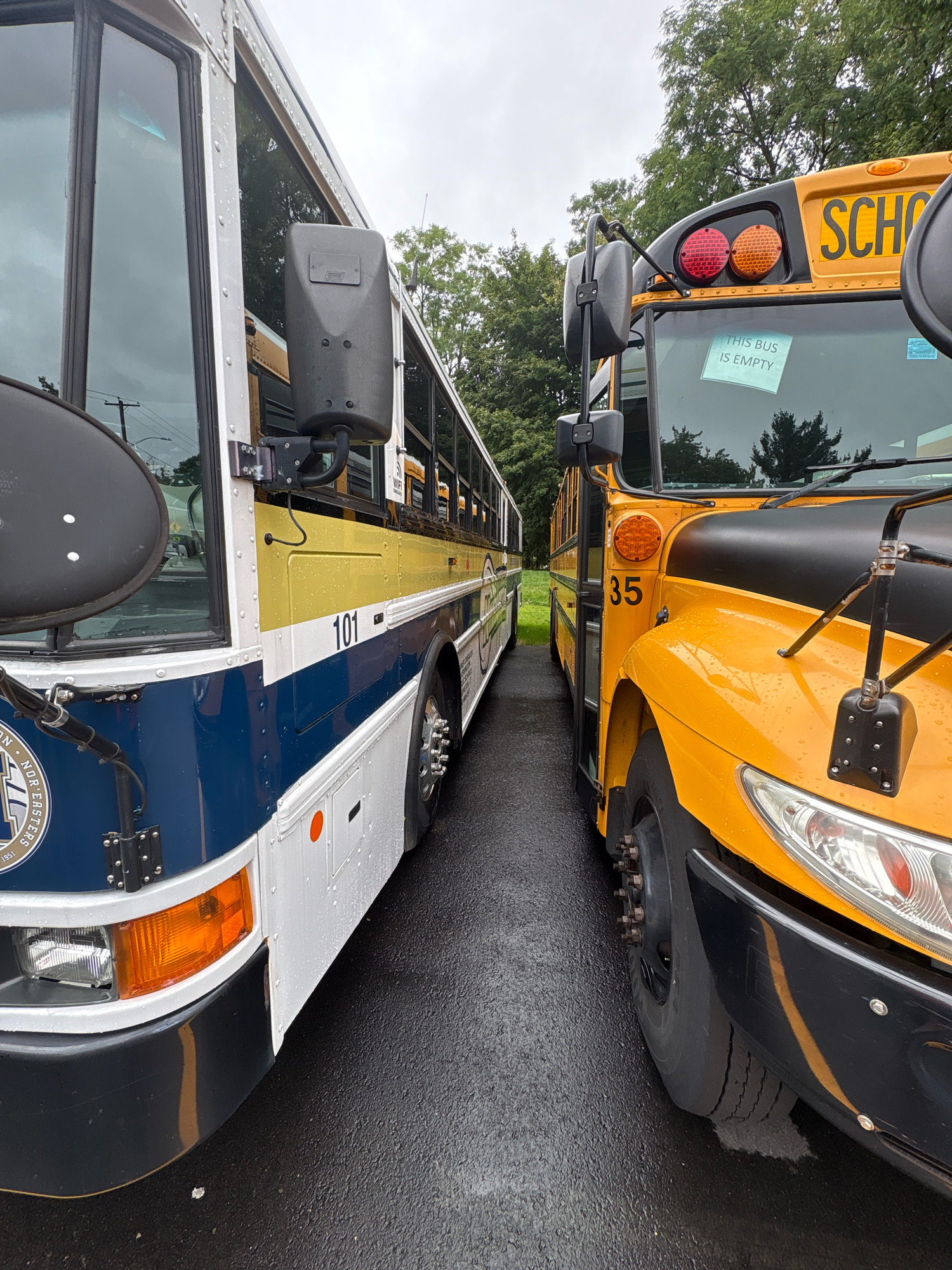 Two school buses are parked side-by-side on a wet asphalt surface, possibly a parking lot. The bus on the left is primarily white with a blue stripe across the bottom and a gold stripe above it. It displays the number "101" prominently. The bus on the right is a standard yellow school bus and shows the number "35" on its side. A sign taped to the windshield of the yellow bus reads "THIS BUS IS EMPTY". The sky is overcast, and trees are visible in the background. There are no people visible in the image. The location is Newington, United States of America.