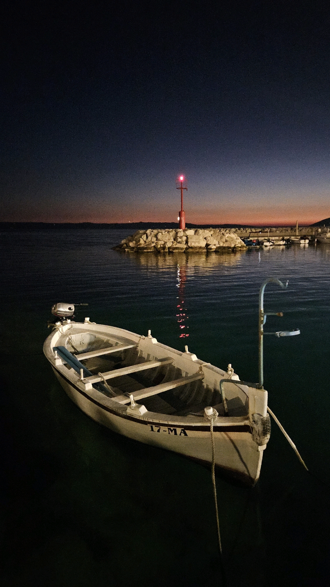 In the city of Općina Tučepi, Croatia, a small white rowboat with an outboard motor is moored in dark, calm water. The boat has a registration number "17-MA" printed on its hull. In the background, a rocky breakwater is illuminated by a red beacon light. Several other boats are docked behind the breakwater. The sky is a deep twilight blue, with faint hints of orange and pink near the horizon, suggesting either dawn or dusk. The scene is lit by artificial lights, casting reflections on the water's surface.