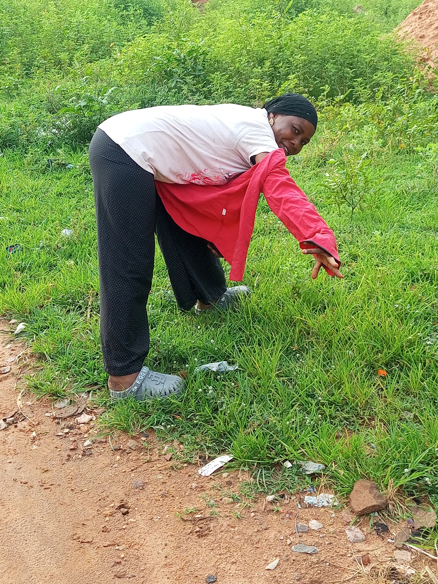 A young Black woman is bending over in a grassy area with some dirt and debris on the ground. She is wearing a white t-shirt and black pants with a white polka dot pattern. A red jacket is draped over her shoulders and down her arms. She is looking back and smiling at the camera. Her right hand is extended towards the grass. She is wearing grey clogs on her feet. The background is filled with lush green vegetation. The scene appears to be in Tempure, Nigeria.