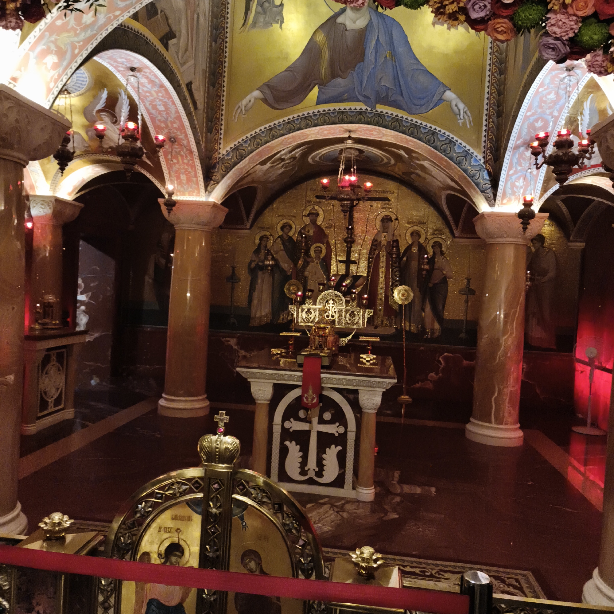 The image depicts the interior of a dimly lit, ornate chapel or crypt, likely in Yekaterinburg, Russia, given the context of historical significance. The architectural style features arched ceilings adorned with frescoes and decorative patterns in shades of pink, gold, and green. Large, marble-like columns, possibly in a reddish-brown hue with white veining, support the arched structures.
The central focus of the scene appears to be an altar or reliquary, adorned with intricate golden metalwork, red fabric, and possibly religious icons. A large golden cross, topped with a smaller cross, is prominently displayed behind the altar. Mosaic artwork depicting religious figures, including a central figure of Christ and surrounding saints, covers the gold-backed walls. Several ornate candelabra with red-lit candles are visible, contributing to the atmospheric lighting.
In the foreground, a golden, crown-like structure with a cross on top is partially visible, suggesting it might be part of an iconostasis or another significant religious artifact. A red ribbon or barrier is positioned in front of this foreground object. The floor is a deep, reflective red marble. The overall ambiance is solemn and rich with religious symbolism.