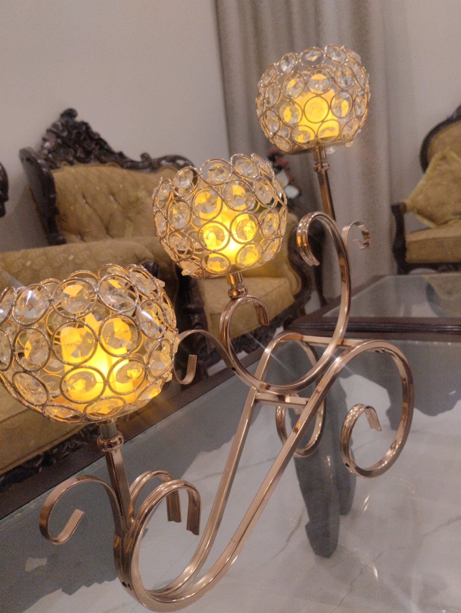 A close-up, eye-level shot features a three-branched golden candelabra placed on a glass table. Each branch holds a spherical candle holder encrusted with clear, multifaceted crystals, through which the warm glow of the lit candles is visible. The candelabra's base is ornate, with swirling metalwork in a matching golden hue.
In the blurred background, two plush armchairs with elaborate wooden frames and golden upholstery suggest an indoor, possibly formal, setting. The lighting is dim and warm, creating a cozy ambiance, and the time of day is indeterminate, but the scene evokes a feeling of tranquility or relaxation. There is no visible text in the image.