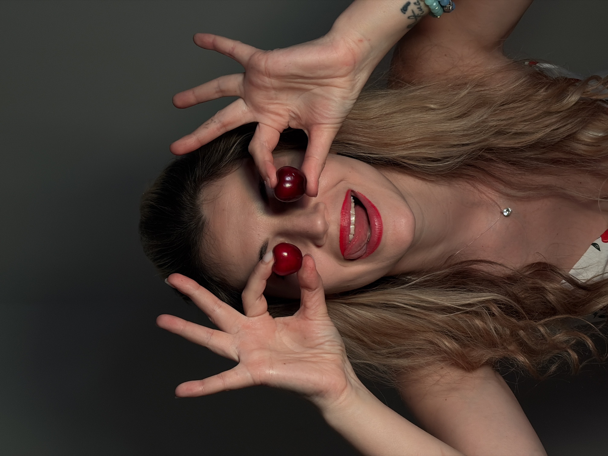 A woman with long, wavy blonde hair is holding cherries in front of her eyes. Her mouth is open, with her tongue sticking out and wearing red lipstick. She has light skin and is wearing a necklace with a small pendant. Her left arm has a tattoo. The background is a dark gray. The location is not discernible.