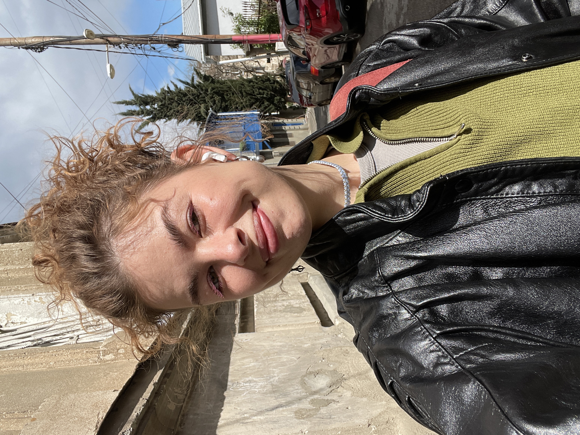 A young woman poses for a selfie outside on a sunny day in Tbilisi, Georgia. She wears a black leather jacket over a green sweater and a silver necklace. Her curly hair is light brown, and she has an earbud in her left ear. Behind her are parked cars, trees, utility poles, and buildings, suggesting a residential street. The ground and a nearby wall appear to be made of concrete.