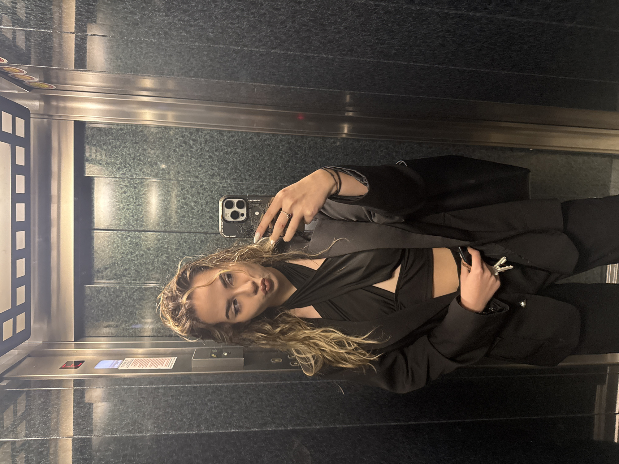 A young woman with blonde, wavy hair is taking a selfie in what appears to be an elevator. She is wearing a black wrap-style top, a black blazer, and black pants. In her right hand, she is holding a set of keys. Her left hand is holding a smartphone to take the photo. The elevator has silver walls and a mirrored panel. There is an elevator control panel with several buttons and an information plaque visible on the left side of the image. No specific landmarks that would indicate the location in Blagoevgrad are visible.