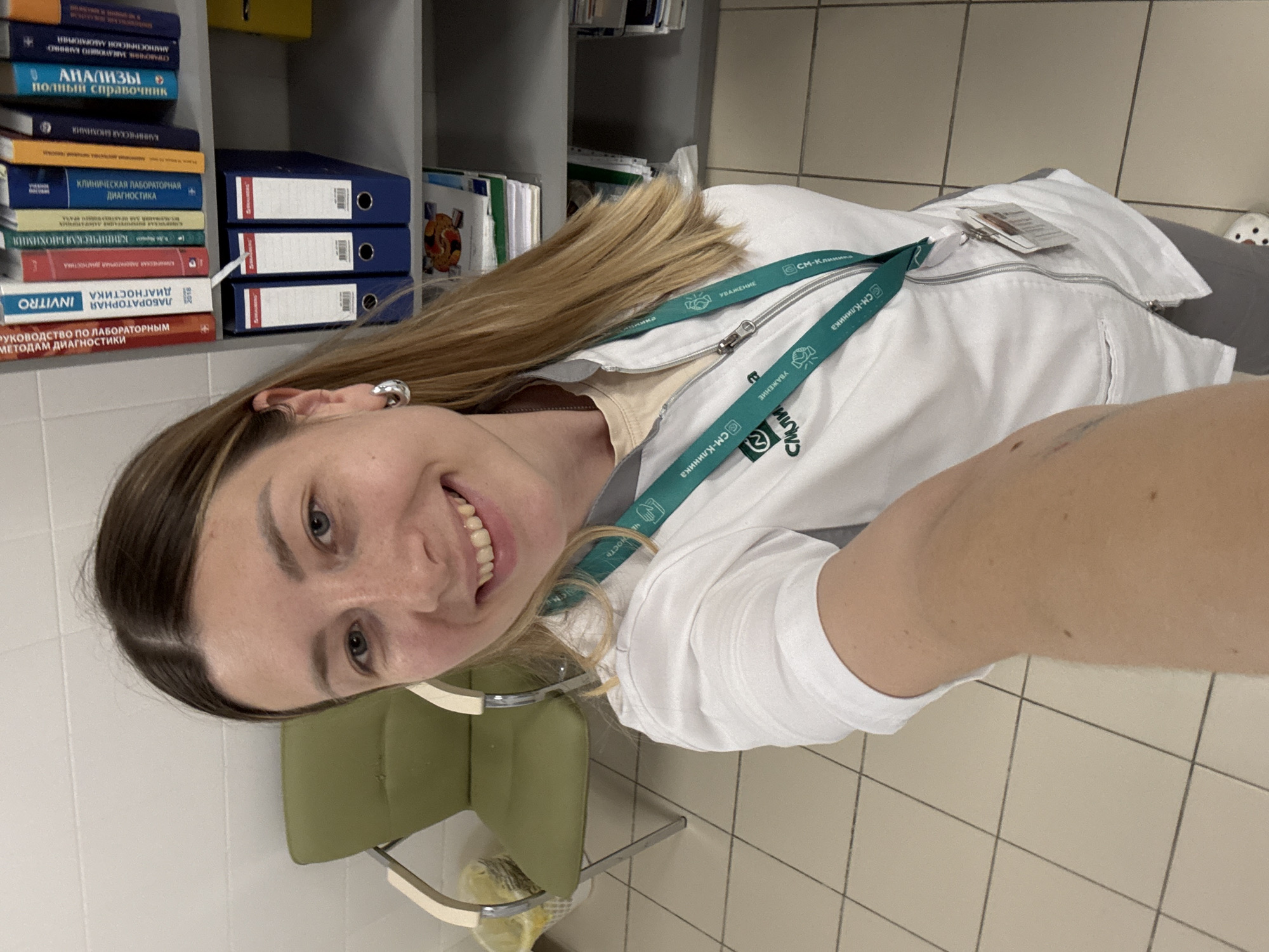 A woman with blonde hair and blue eyes, wearing a white lab coat with a green lanyard, smiles at the camera. She is standing in what appears to be a medical office or clinic, with shelves of books and binders in the background. The room has tiled flooring and a green chair is visible to the left of the woman. The overall impression is that of a professional setting.