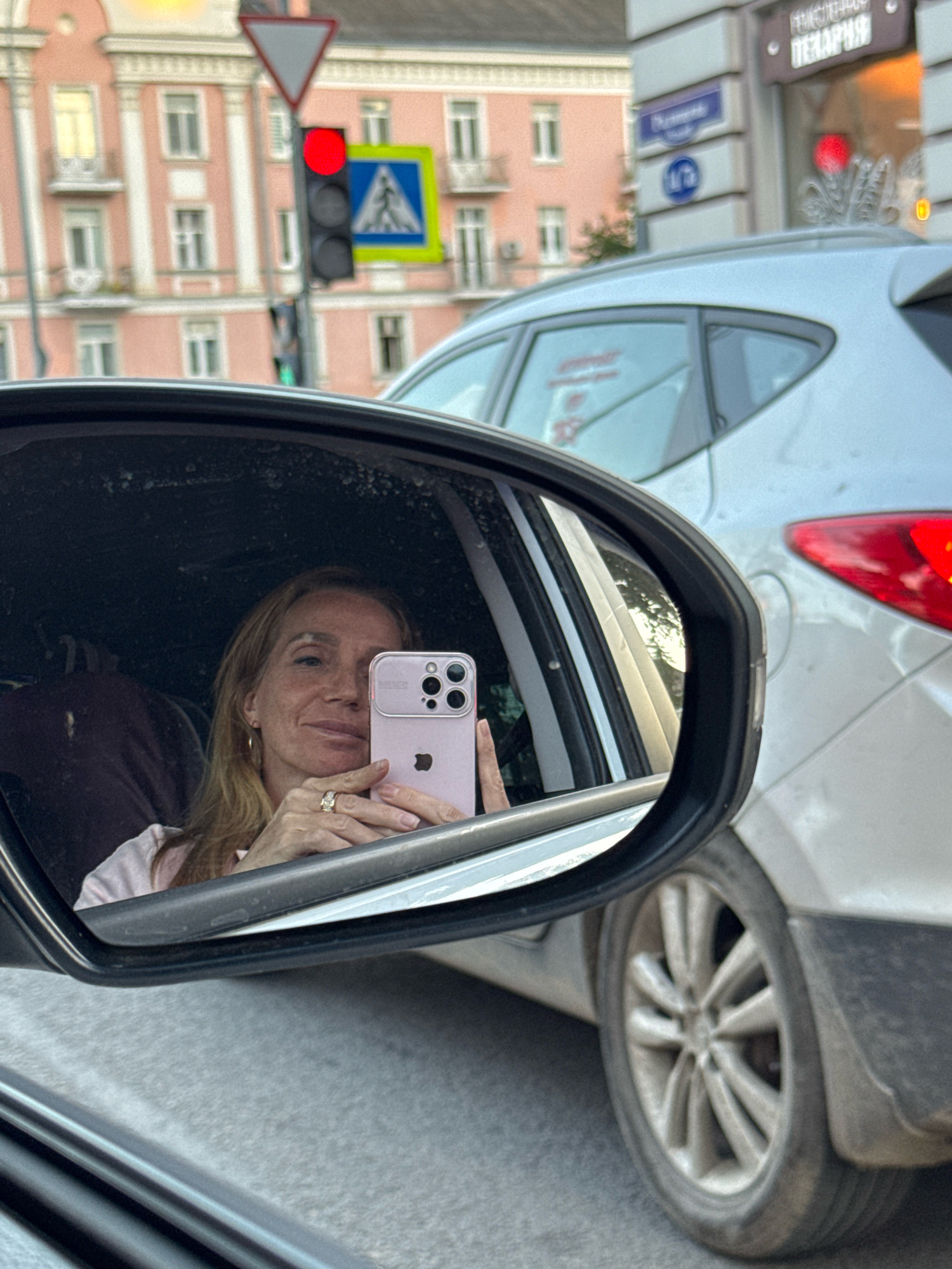 A woman is taking a selfie in a car's side-view mirror. She is looking at her phone, which she is holding in her right hand. She has blonde hair and is wearing a pink shirt and a ring on her left hand. The car is silver and is in traffic. The background shows buildings, traffic lights, and a pedestrian crossing sign. The woman appears to be in a city. This image was taken in Tver, Russia. Specifically, it was taken on Oktyabrsky Prospekt, near the intersection with Sverdlova Street. The red traffic light indicates that the car is stopped.