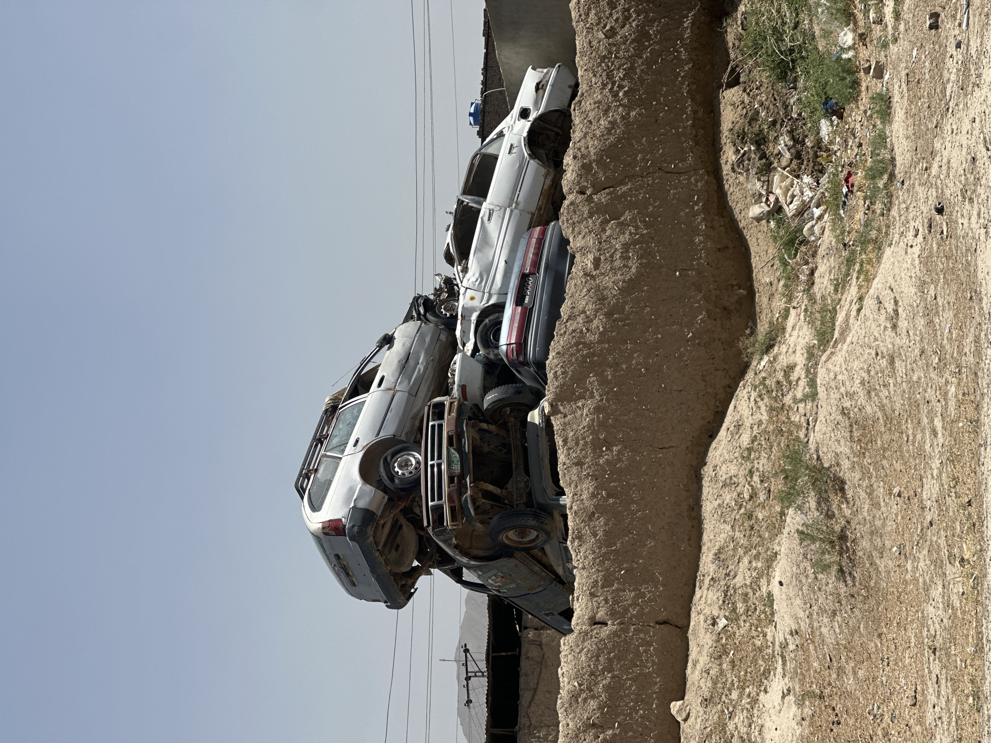 In Kanozai, Pakistan, a junkyard is piled high with several wrecked cars. In the foreground, a mud-brick wall with visible cracks separates the junkyard from a dry, dusty area. Sparse green vegetation and debris are scattered across the ground. In the background, a light blue sky stretches over the scene, with distant mountains or hills visible on the horizon. The stacked cars are the central focus, appearing heavily damaged and weathered.