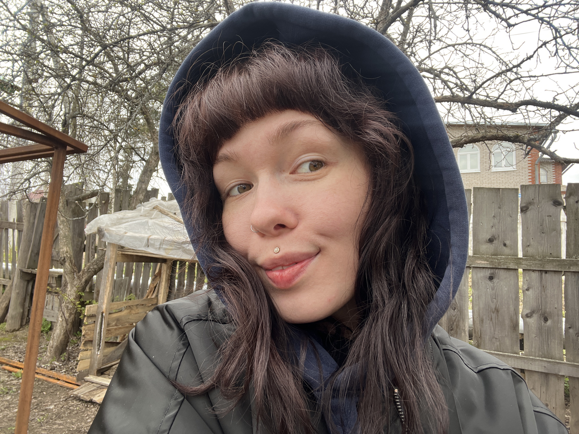 A person with brown hair and a hooded jacket poses for a photo outdoors. The subject has a nose ring and a lip piercing. Behind the person, there is a wooden fence, trees without leaves, and what appears to be a wooden structure covered in plastic. A brick building is visible in the distance. This image was taken in Chaikovsky, Russia.