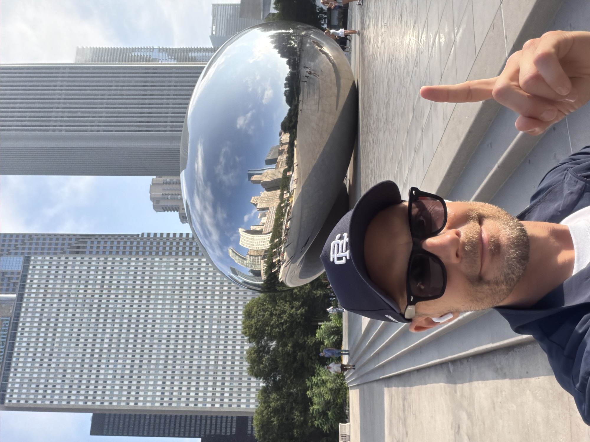The image shows a man taking a selfie in front of "Cloud Gate," also known as "The Bean," in Millennium Park, Chicago, Illinois, United States of America. The man is wearing a dark baseball cap, sunglasses, and a dark jacket over a white t-shirt. He has a beard and is pointing his index finger upwards. In the background, "The Bean" reflects the surrounding city skyline, including tall skyscrapers. The sculpture is a large, highly polished, bean-shaped object made of stainless steel. Steps are visible in the foreground leading down to the plaza area where the sculpture is located. Several other people are visible in the background, some walking and others interacting with the sculpture. The sky is bright and appears to be daytime.