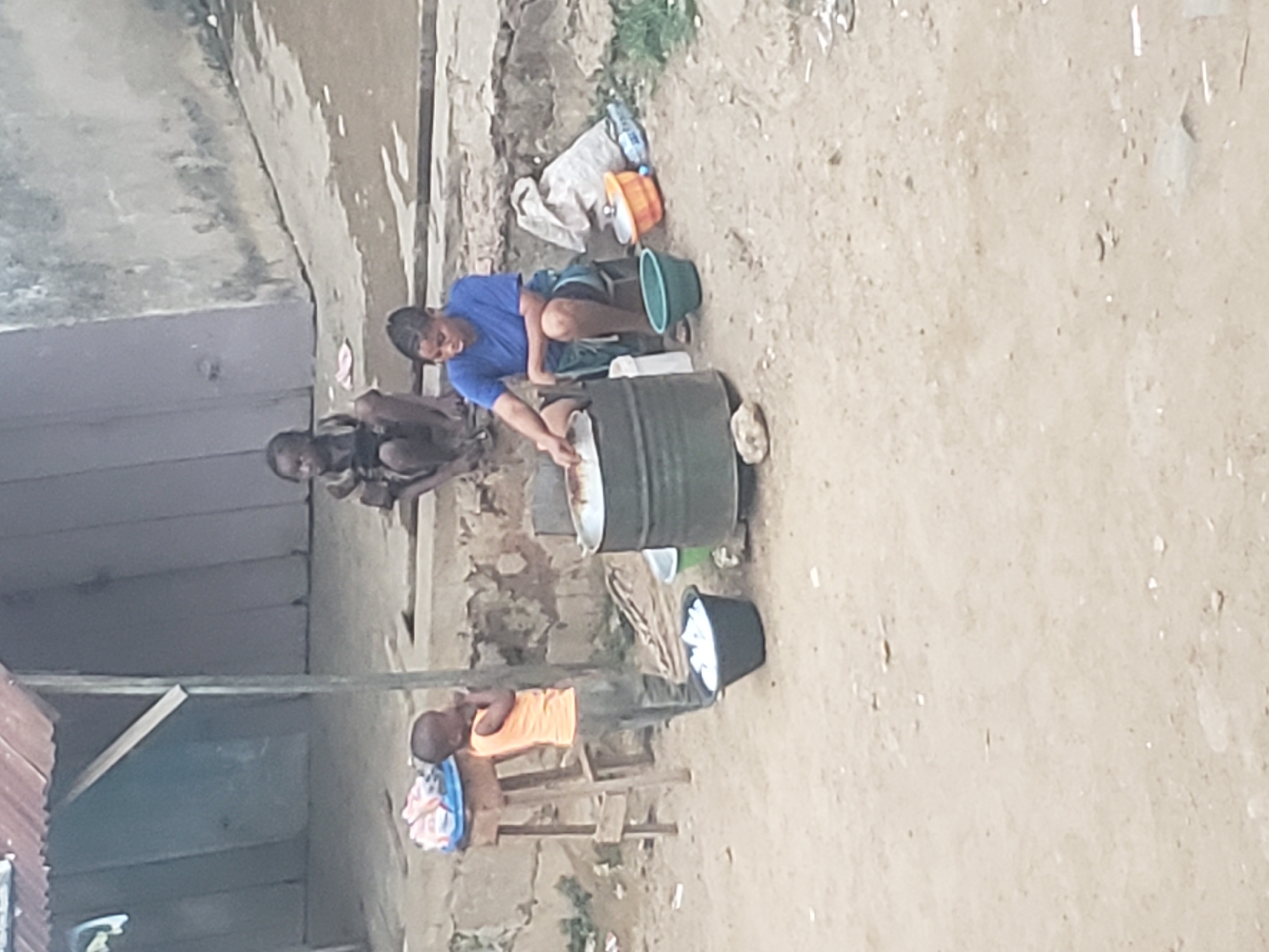 In the city of Alalubosa, Nigeria, a young person wearing a blue shirt is bending over a metal drum, seemingly washing clothes. Beside them, another individual sits on the ground. Nearby, an individual sits on a wooden stool holding a blue container. The ground is sandy and bare. In the background, there is a building with a grey wall and a corrugated metal roof. There are containers, including buckets and bags, strewn around.