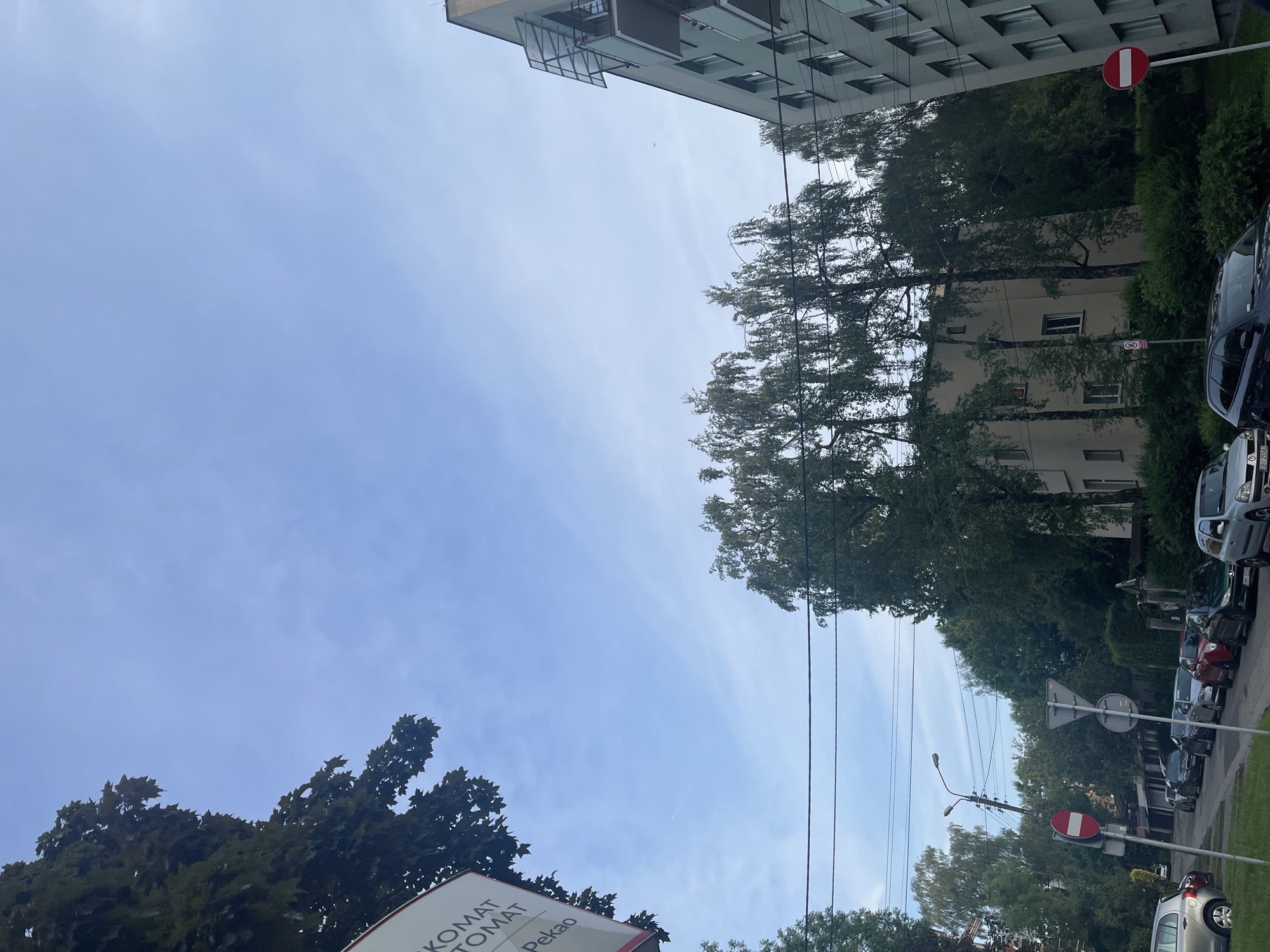 This image captures an urban streetscape on a clear day, viewed from a slightly upward angle, with the original orientation being portrait but appearing landscape when displayed. The scene is dominated by a bright blue sky with subtle, wispy clouds, suggesting fair weather during daytime.
On the left side of the frame (when reoriented), a modern, light-colored multi-story building with numerous windows and balconies is visible. On the right, a street runs parallel to the viewer, lined with parked cars and a variety of green trees and hedges, some of which are tall and slender. Behind these trees, another, possibly older, building can be glimpsed.
Several street signs and utility poles with wires are present along the street. A prominent red circular "No Entry" sign with a white horizontal bar is visible on the right, with another, smaller sign (possibly indicating parking restrictions) beneath it. Further down the street, a triangular yield sign is also visible. Utility poles support overhead lines crisscrossing the sky.
In the lower left corner, a large white sign is partially visible. The text on this sign includes "KOMAT" (likely part of "BANKOMAT"), "TOMAT", and "Pekao". Given that "Pekao" is a Polish bank, this sign is likely advertising a "Bankomat Pekao" (Pekao ATM), suggesting the location is in Poland. No people are visible in the scene. The overall impression is that of a quiet, everyday urban environment.