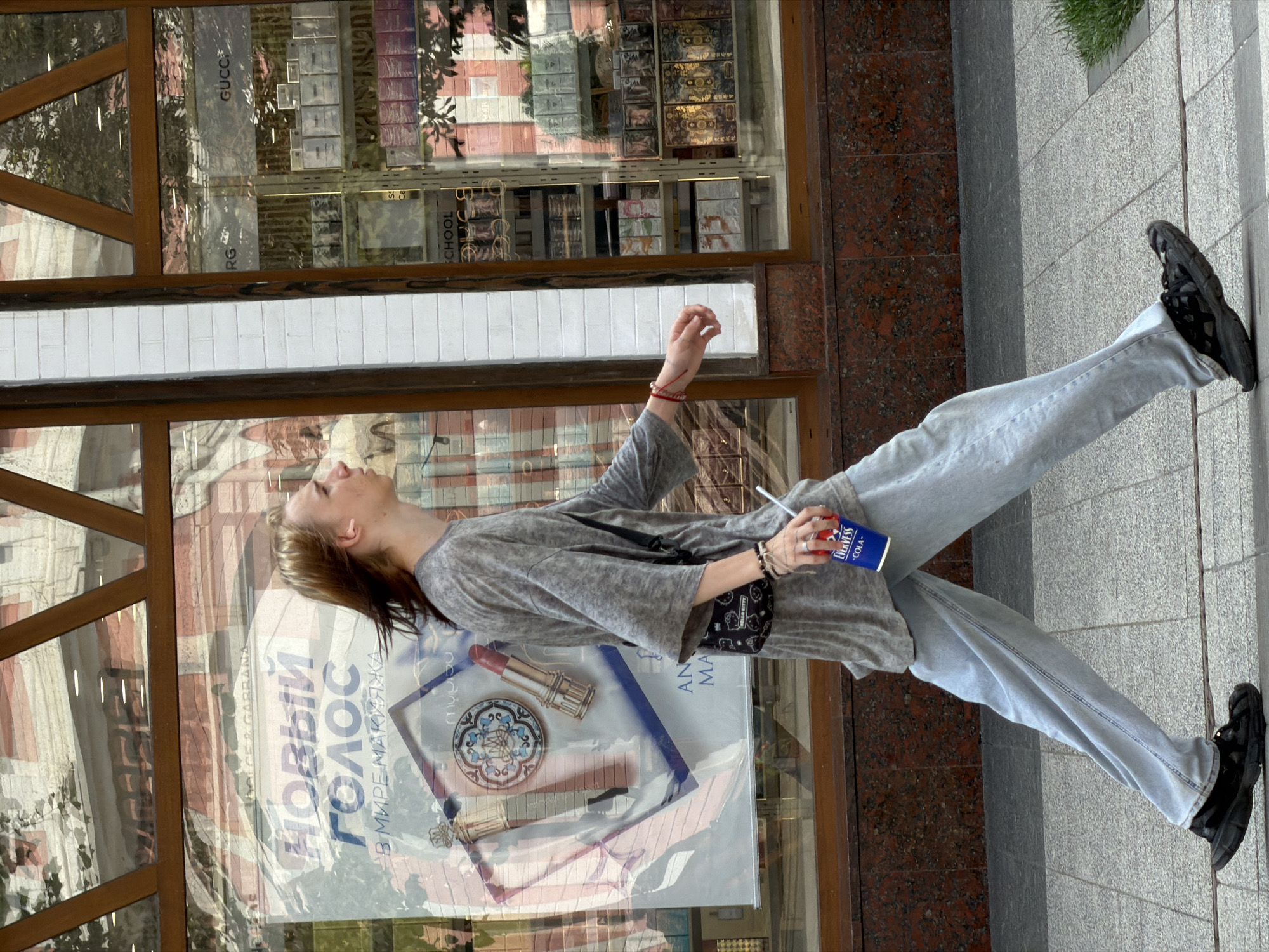 A young person with long, light brown hair walks on a sidewalk in front of a storefront. The person is wearing a loose, grey, short-sleeved shirt, light blue jeans, and black, chunky sneakers. They are carrying a blue cup with a white straw in their right hand and have a small, black purse with a white Hello Kitty pattern slung across their chest. A red string bracelet is visible on their left wrist. The storefront window displays advertisements and rows of perfume or cosmetic products. The street is paved with grey rectangular tiles, and a small patch of green grass is visible to the right. The scene appears to be in a city environment.