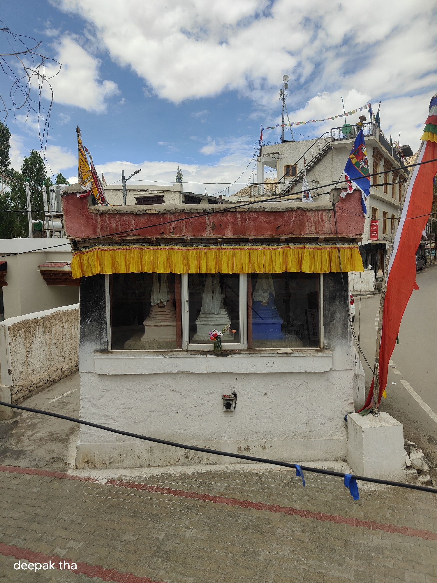 The image depicts a small, white, stucco building with a red-painted facade and a yellow fabric valance above a large glass window. Inside the window, three white stupas are visible. The building is situated in the city of Leh, India, as indicated by the name "deepak tha" in the lower left corner. In the background, a multi-story building with a balcony and stairs can be seen, adorned with prayer flags and other colorful flags. A large red and white flag is prominently displayed on the right side of the image. The street in front of the building is paved with interlocking concrete blocks and features a white dividing line. The sky is partly cloudy.