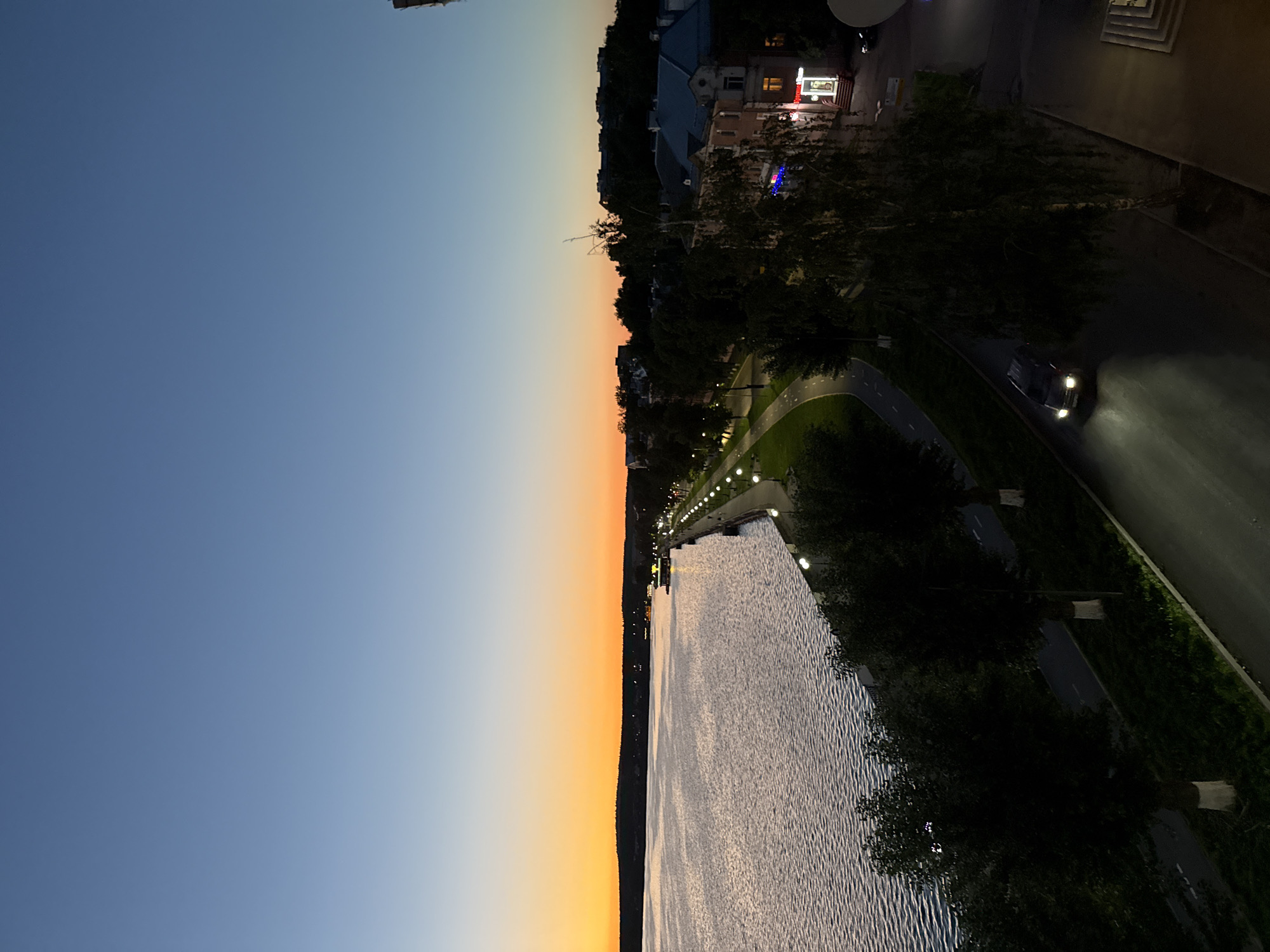 The image depicts a twilight scene along the Votkinsk Reservoir embankment in Votkinsk, Russia. The sky transitions from a pale blue near the zenith to a vibrant orange and yellow glow at the horizon, indicating a sunset or sunrise. The reservoir, with its rippling surface reflecting the sky, occupies the left side of the frame.
Along the embankment, a paved walkway lined with lampposts follows the curve of the water. Adjacent to the walkway is a green lawn and a path, likely for cycling or recreation. Trees are planted along the embankment, some with their trunks whitewashed, a common practice in Russia.
Further inland, buildings with tiled roofs are visible, illuminated by streetlights and interior lights. One building appears to be a shop or cafe with illuminated signage. A dark-colored car with its headlights on is driving on a road that runs parallel to the embankment, its headlights casting a bright beam onto the asphalt. The overall atmosphere is tranquil, with the artificial lights of the city beginning to dominate as natural light fades.