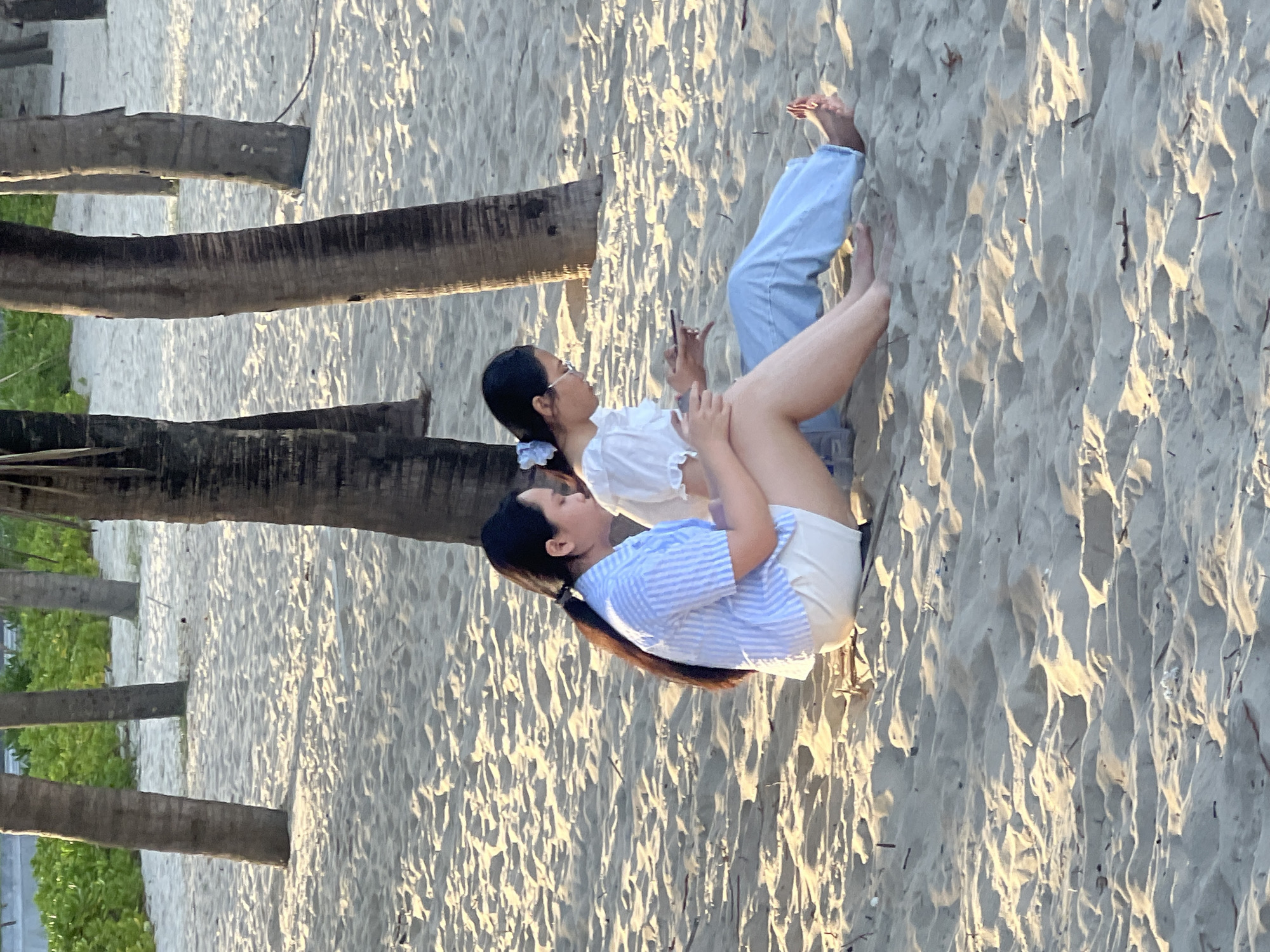 Two young women are sitting on a sandy beach in Đà Nẵng, Vietnam, amidst palm trees. The woman on the left is wearing a blue and white striped short-sleeved shirt and white shorts, with her dark hair tied in a ponytail. The woman on the right is wearing a white ruffled top and light blue pants, with glasses and her dark hair pulled back with a light blue scrunchie. Both are looking down at their phones, with the woman on the left holding her phone in her left hand and the woman on the right holding hers in her right hand. The sand is light beige, with ripples and shadows from the surrounding palm trees casting patterns on the ground. In the background, lush green foliage is visible behind the palm trees. The lighting suggests it is late afternoon or early morning.