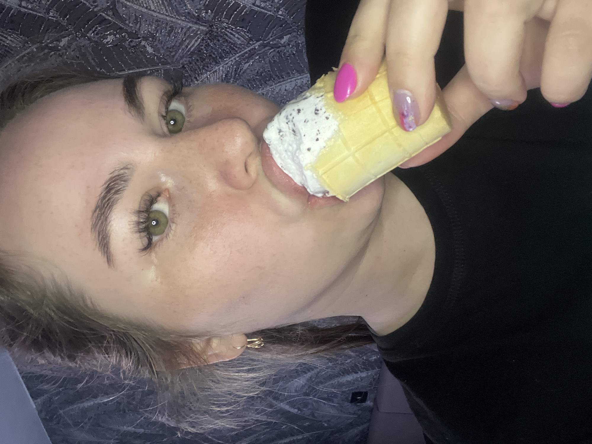 A fair-skinned female is shown in a close-up, angled view, eating an ice cream cone. The cone has a waffle pattern and appears to be filled with cookies and cream flavored ice cream. Her nails are painted with a variety of colors, including pink and a floral pattern. She is wearing a black shirt. Her hair is light brown and a gold earring is visible in her left ear. The background appears to be a grey patterned textile. The location in Chaikovsky, Russia, cannot be determined from this image.
