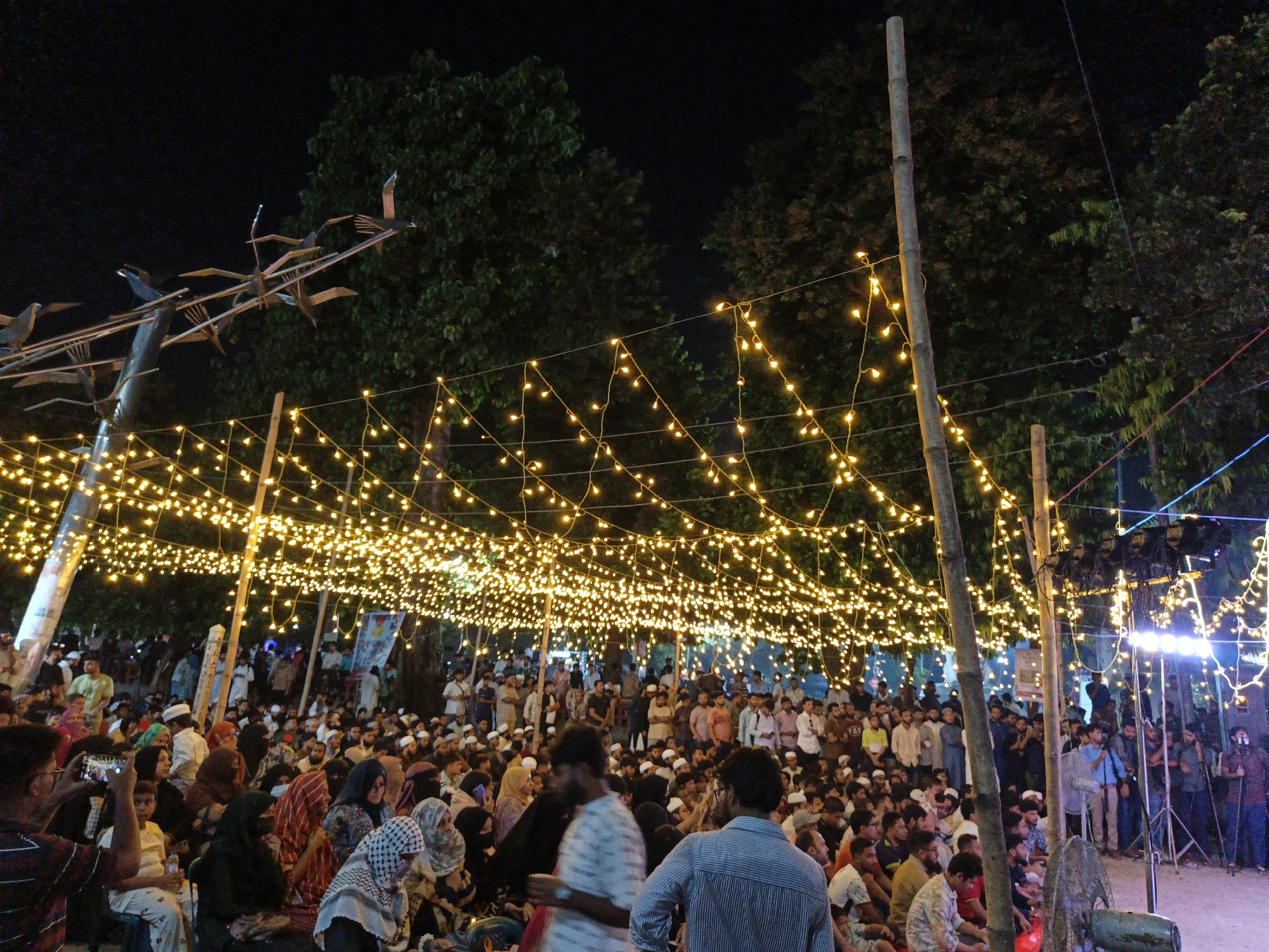 The image depicts a nighttime outdoor gathering of a large crowd, densely packed in an open area. Numerous strings of small, warm-toned lights are suspended overhead, creating a canopy of illumination. Several tall, thin bamboo poles are positioned throughout the scene, supporting the light strings. In the background, dark silhouettes of trees are visible against the night sky. The crowd is diverse, with individuals of various ages dressed in a mix of traditional and modern attire, many wearing head coverings. Some people are seated on the ground, while others stand. One individual in the foreground on the left is holding up a mobile phone to take a photograph. The overall atmosphere appears to be festive or commemorative, suggested by the elaborate lighting and the significant assembly of people. The scene is consistent with public events or celebrations held in urban spaces in Dhaka, Bangladesh, during the evening.