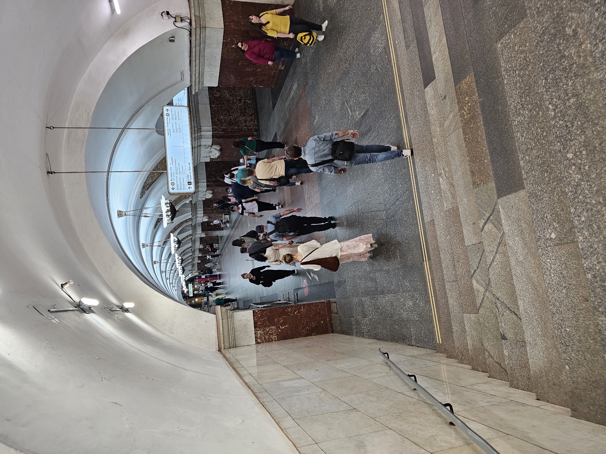 The image shows an underground train station in Moscow, Russia. The station features an arched ceiling, tiled walls, and marble-like details. A staircase with a metal handrail leads down to the platform. Several people are walking through the station. There are signs with station information hanging from the ceiling. The overall lighting is bright.